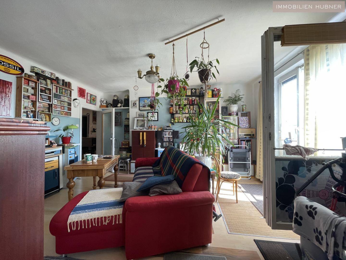 Helles Wohnzimmer mit rotem Sofa, vielen Pflanzen und einem Blick auf den Außenbereich.