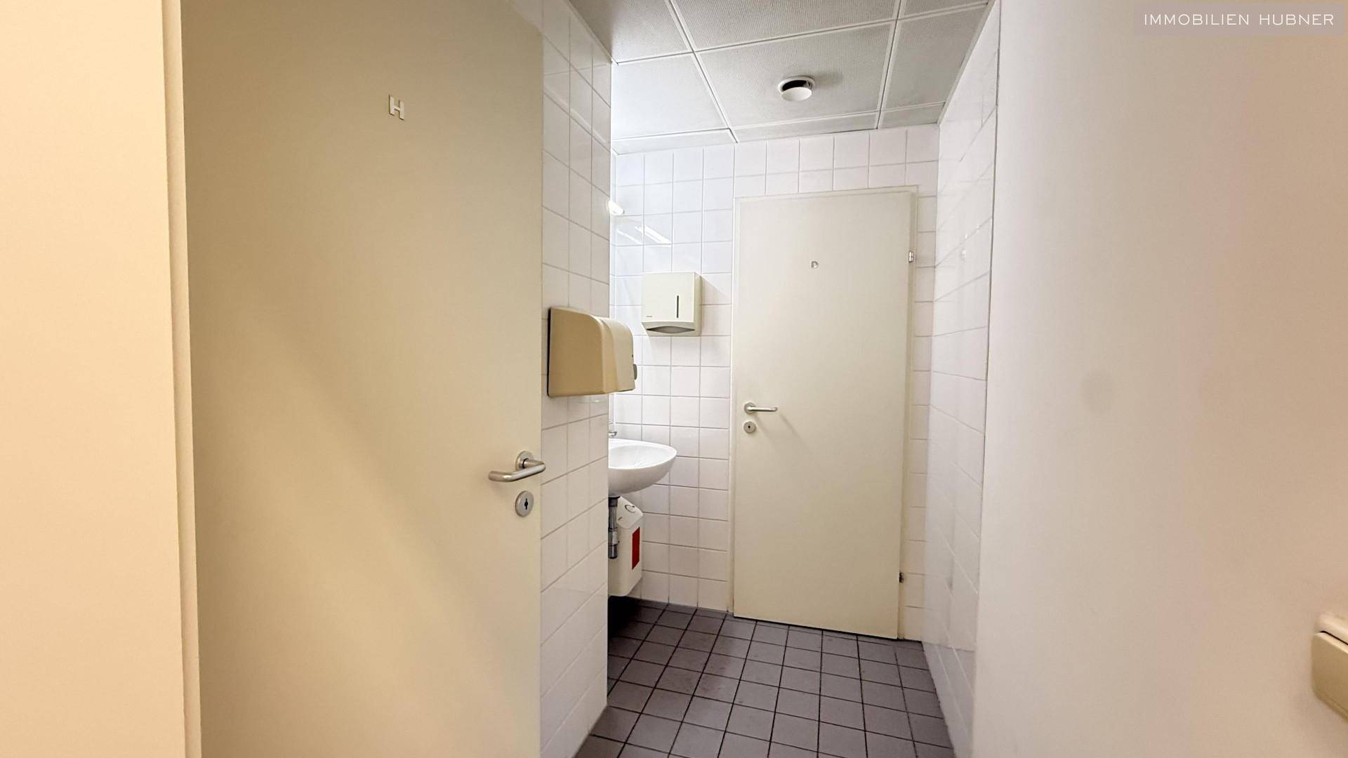 Kleiner Toilettenraum mit weißen Fliesen, Waschbecken und zwei Türen.