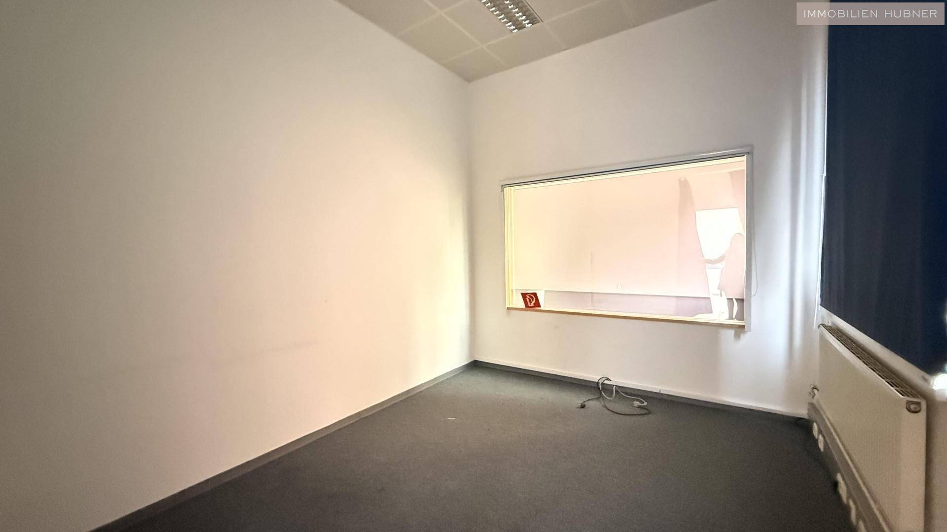 Büro mit Teppichboden, weißen Wänden und einem internen Fenster zu einem Nebenraum.
