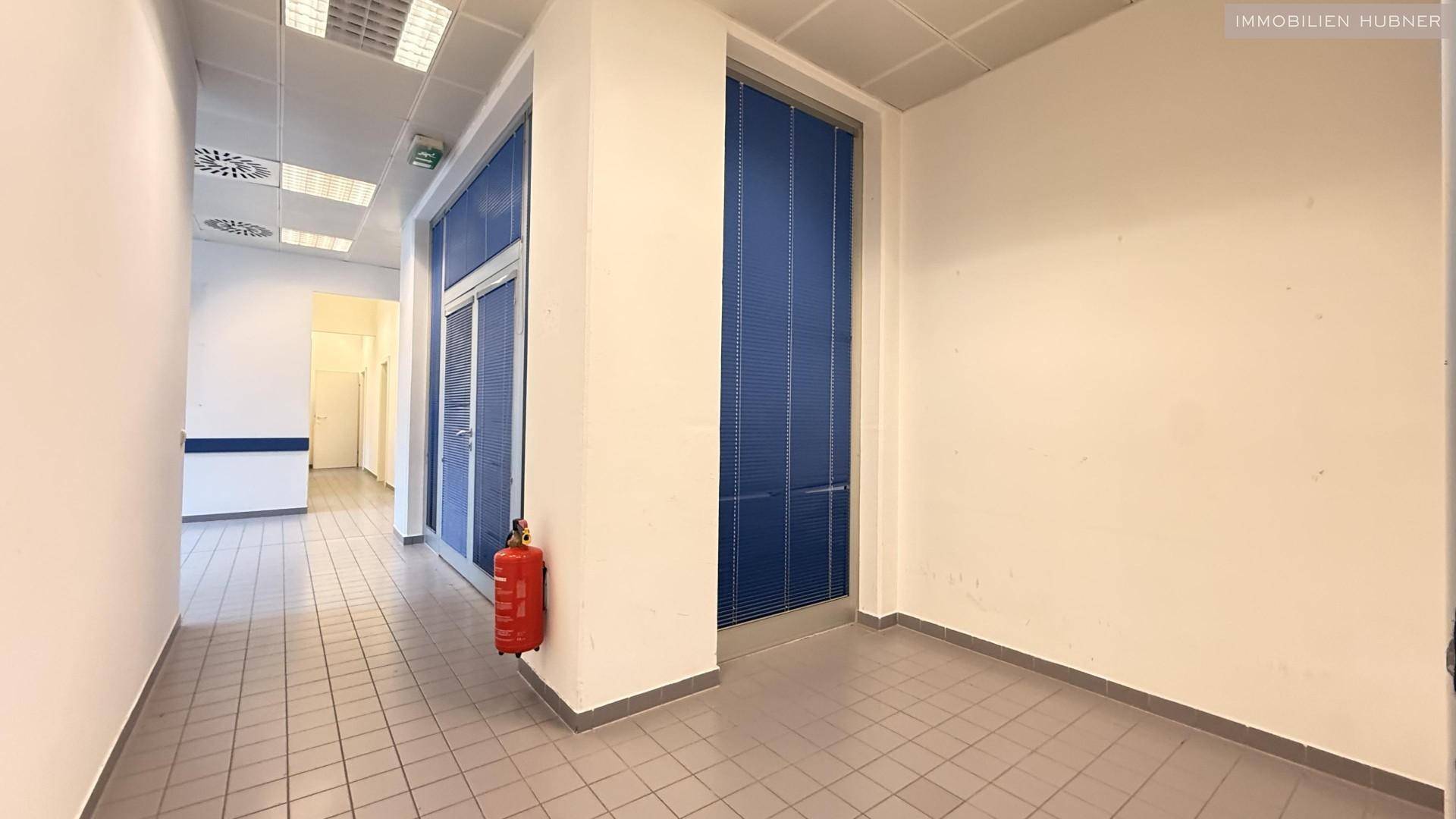 Langer Flur mit Fliesenboden, weißen Wänden und einem Feuerlöscher an der Wand.
