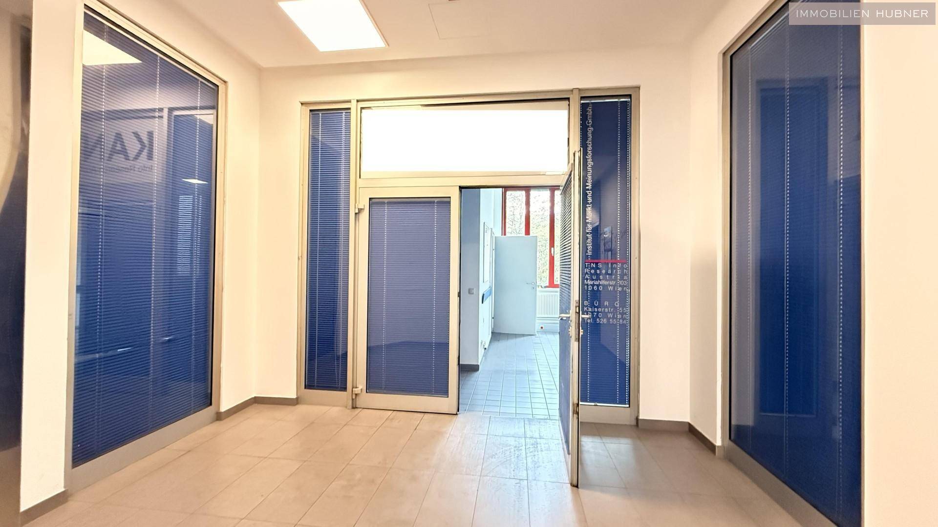 Heller Eingangsbereich mit Fliesenboden und blauen Jalousien an den Glaswänden.