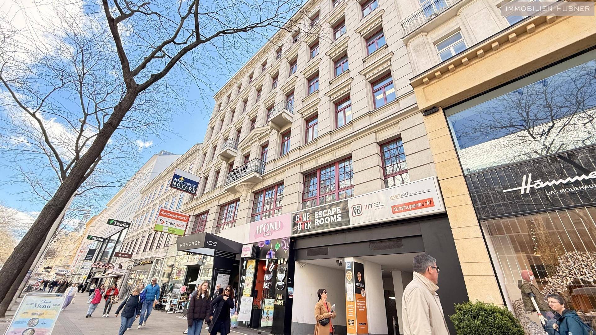 Gebäudeansicht von der Straße mit Passanten, Geschäften und Bäumen im Vordergrund.