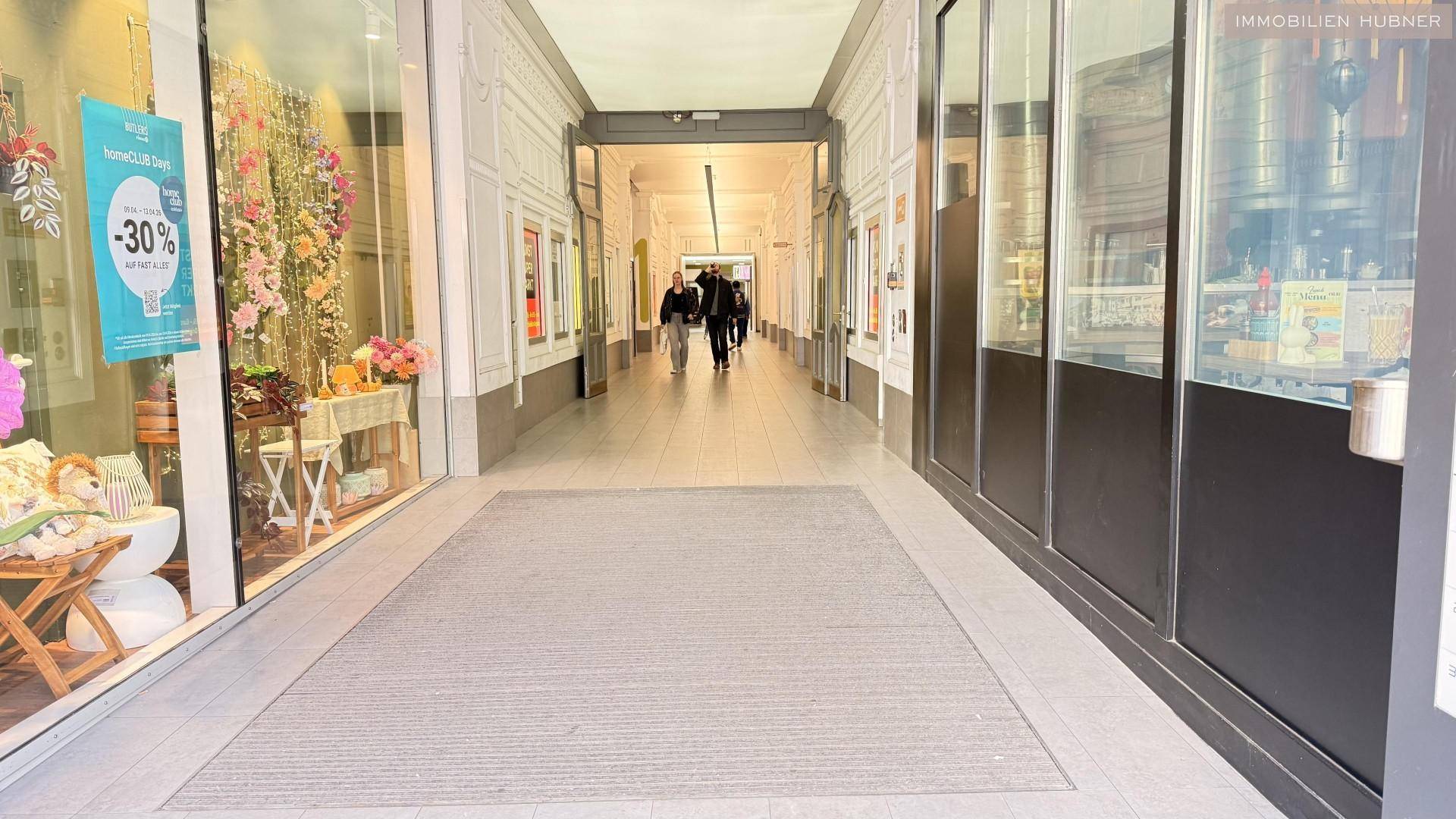 Helle Ladenpassage mit Schaufenstern und Menschen, die durch den Gang gehen.