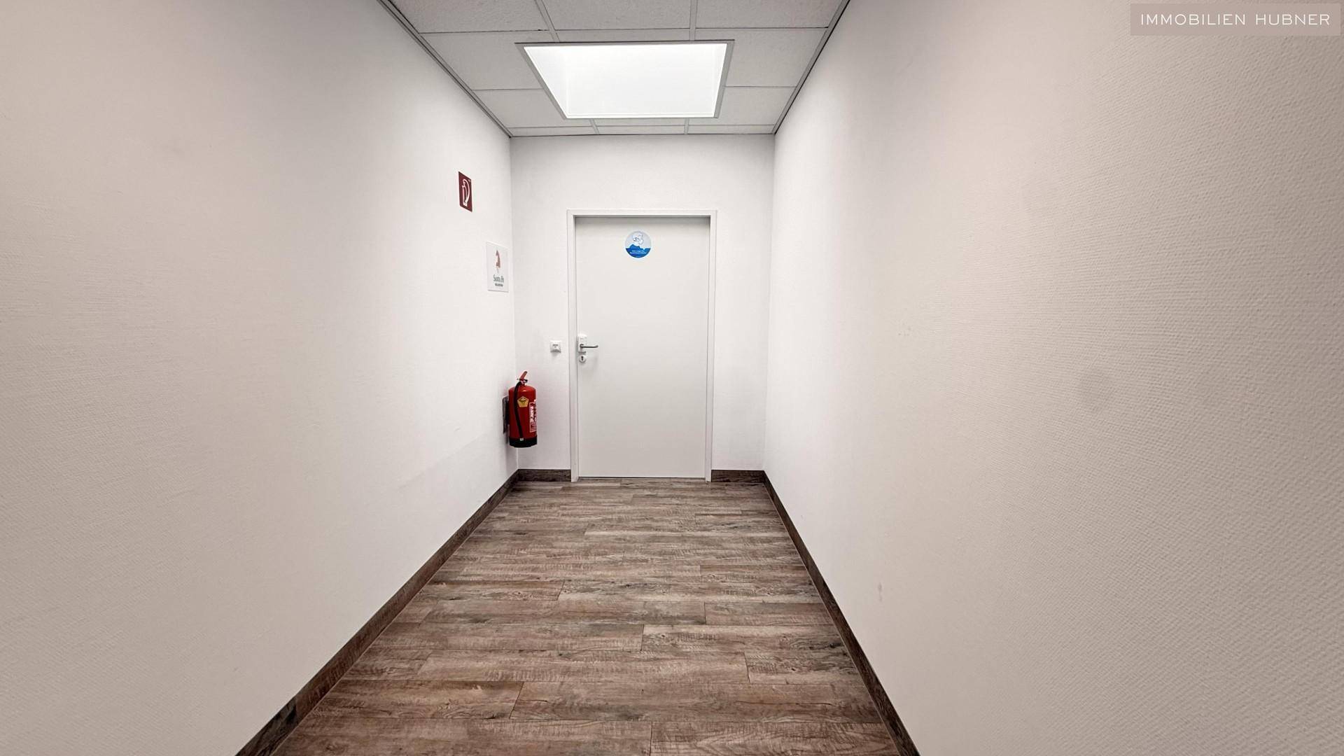 Heller Flur mit Holzoptik-Boden, weiß gestrichenen Wänden und einem Feuerlöscher.