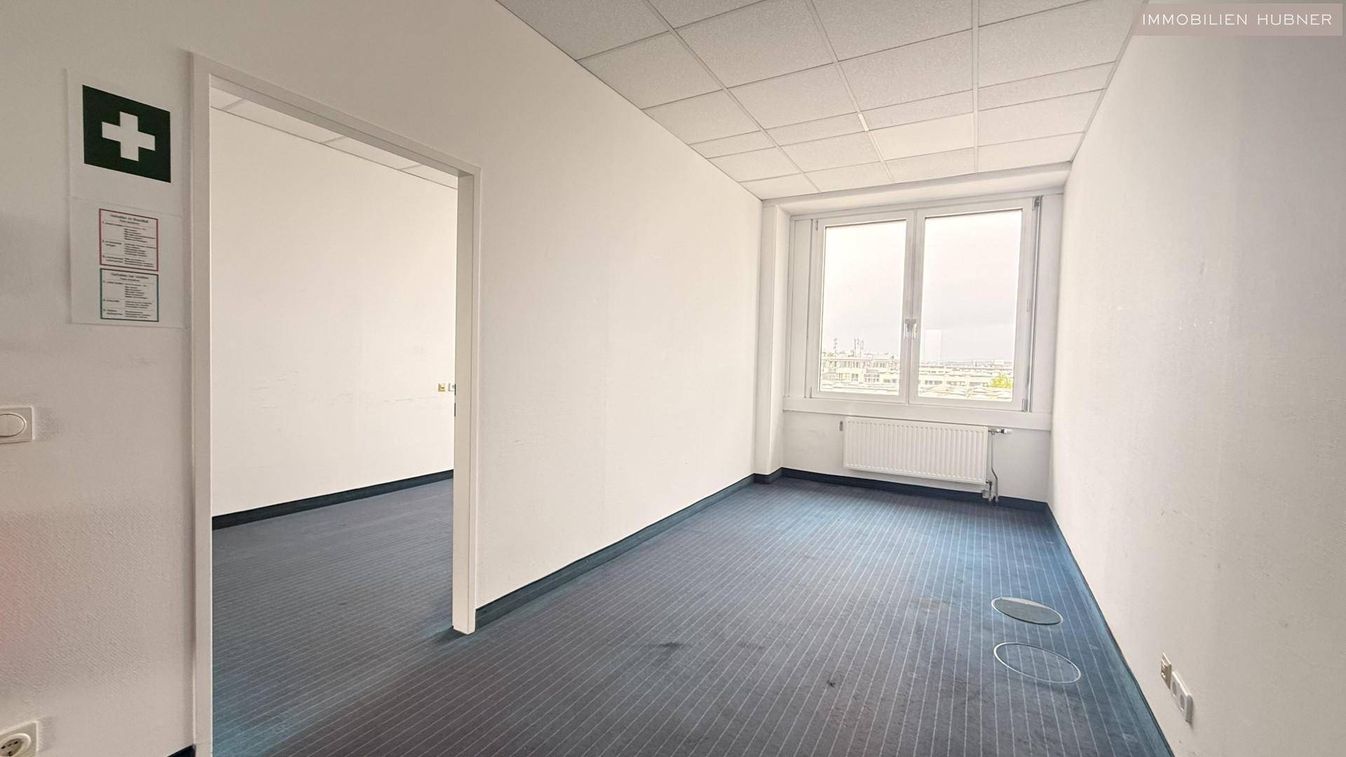 Leeres Büro mit Teppichboden und Fenster, daneben ein Durchgang zu einem weiteren Raum.