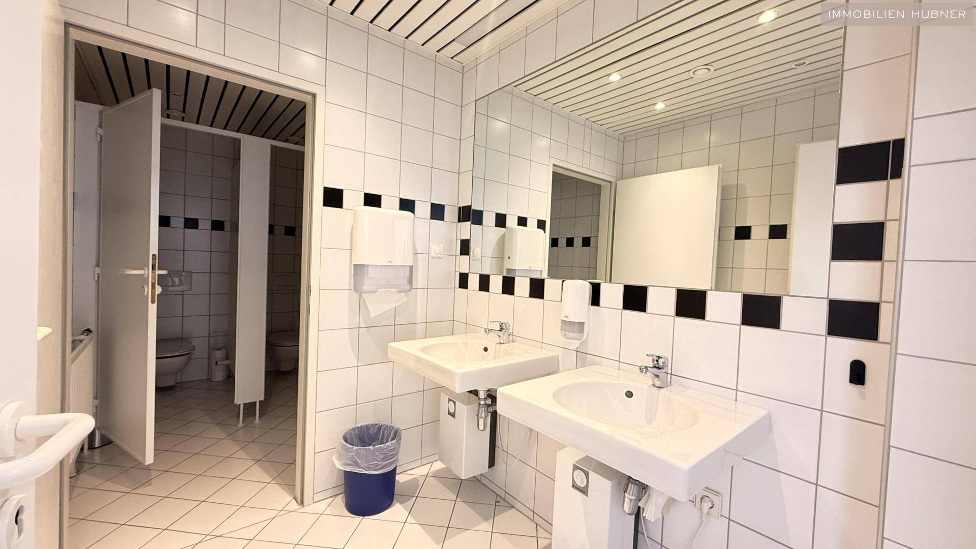 Helle öffentliche Toilettenanlage mit weißen Fliesen, zwei Waschbecken und großen Spiegeln.