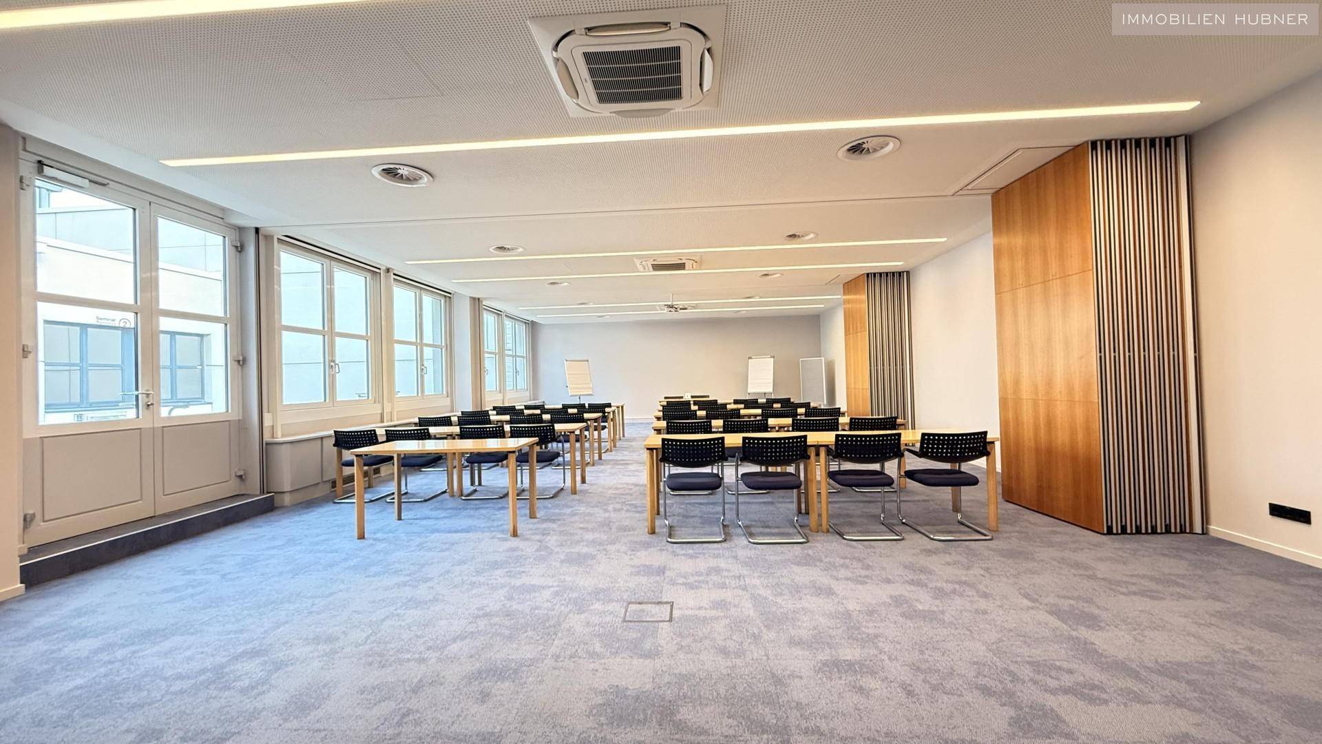 Großer Konferenzraum mit Tischen und Stühlen, großen Fenstern und Teppichboden, bereit für Meetings.