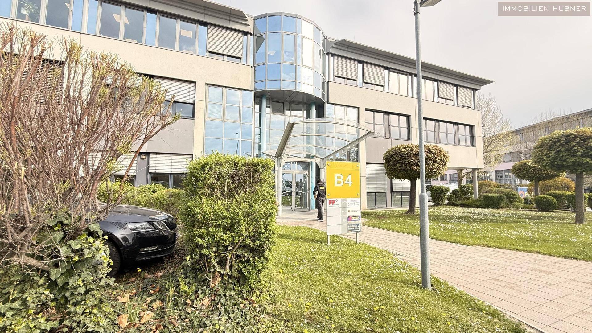 Moderner Büroeingang mit Glasfassade, gepflegter Grünanlage und Beschilderung für Gebäude B4.