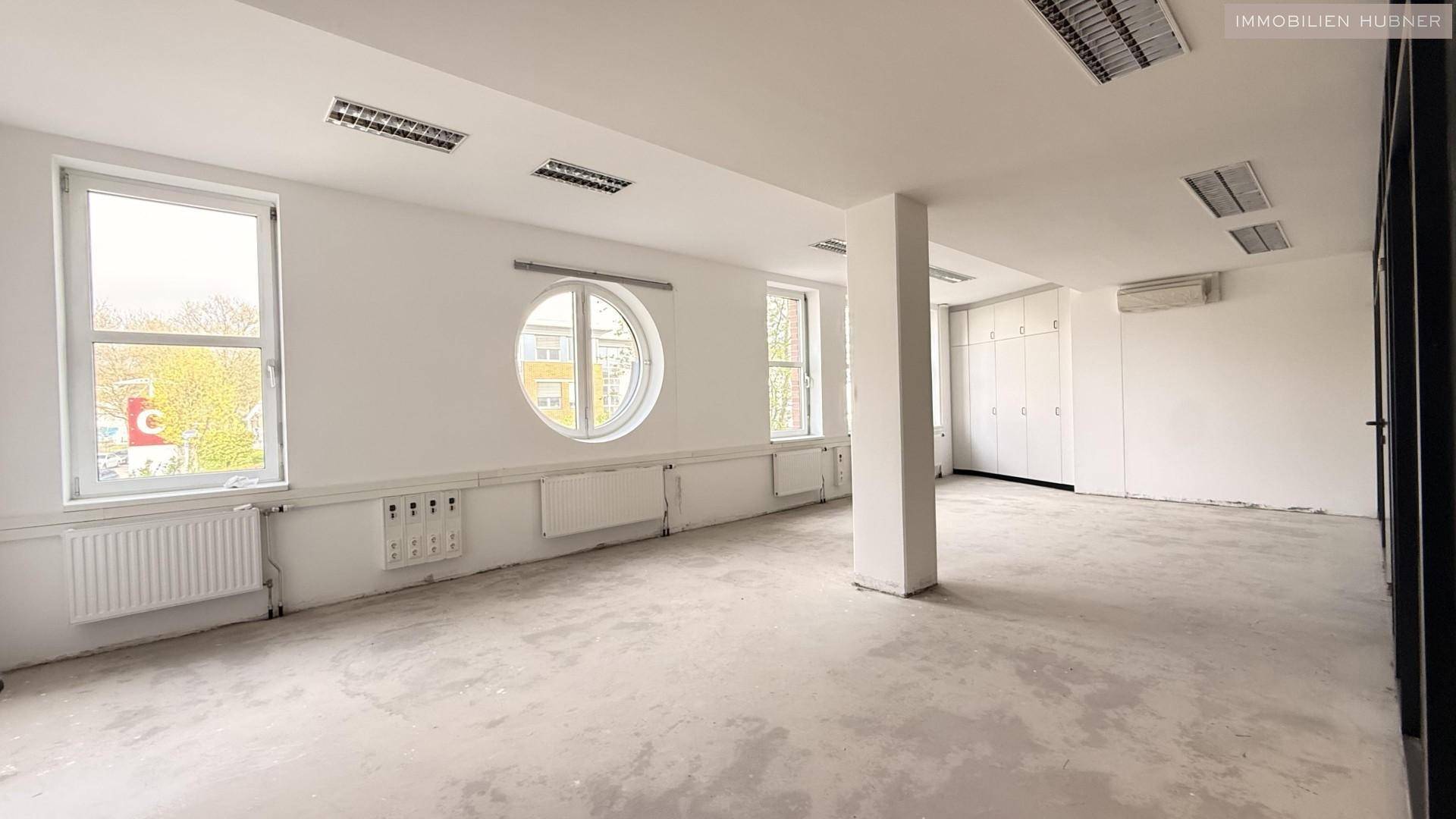 Leeres Büro mit großen Fenstern, darunter ein markantes Rundfenster, und Einbauschränken.