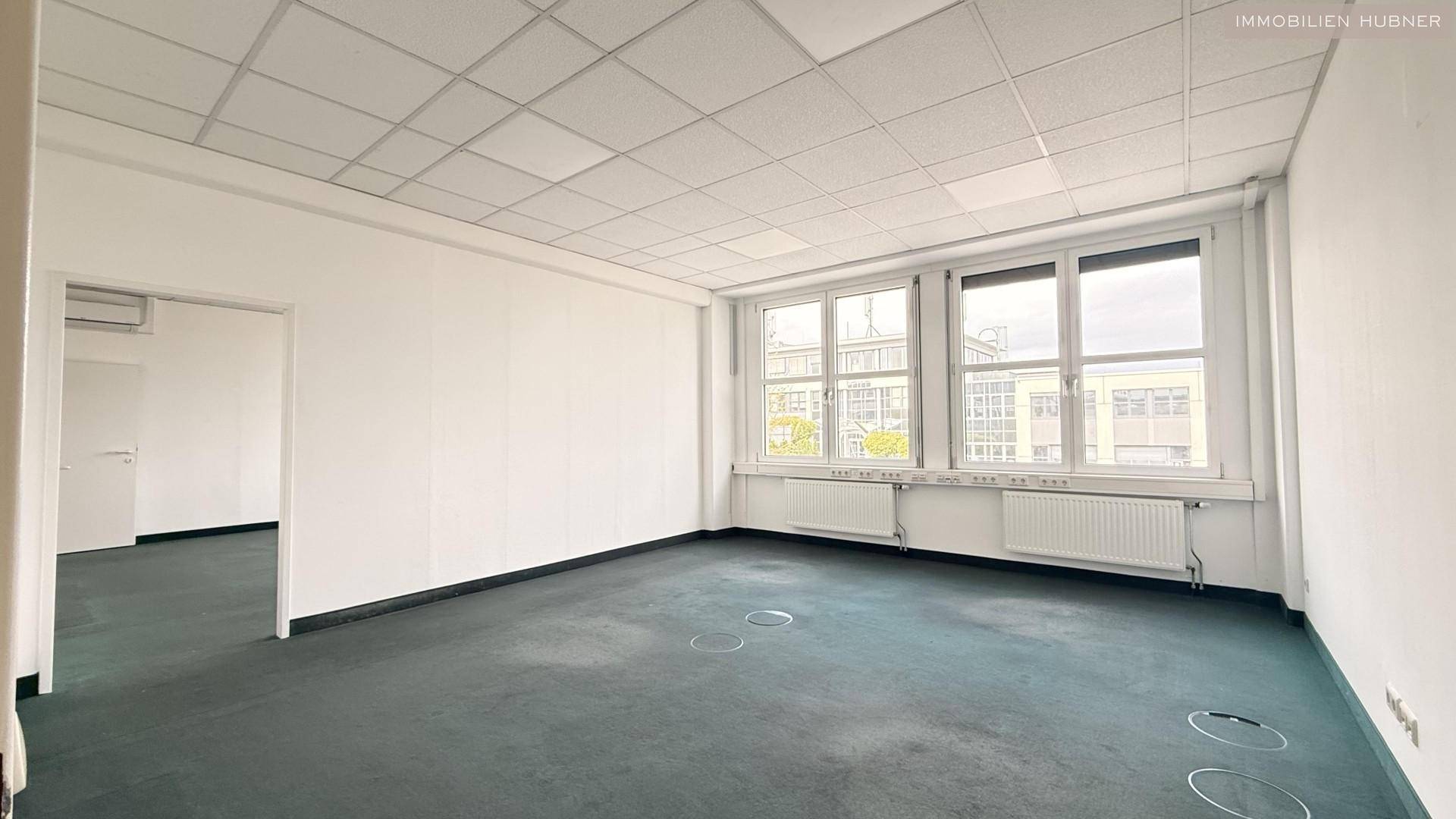 Weitläufiges Büro mit Teppichboden und großen Fenstern, bietet viel Platz und Helligkeit für verschiedene Arbeitsbereiche.