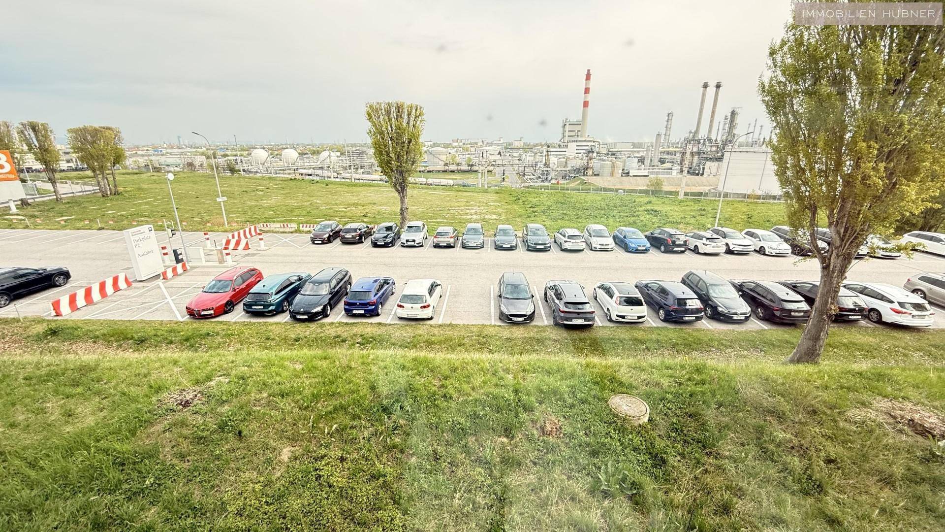 Großer Parkplatz vor einem Industriegebiet mit zahlreichen Fahrzeugen und Bäumen unter bewölktem Himmel.
