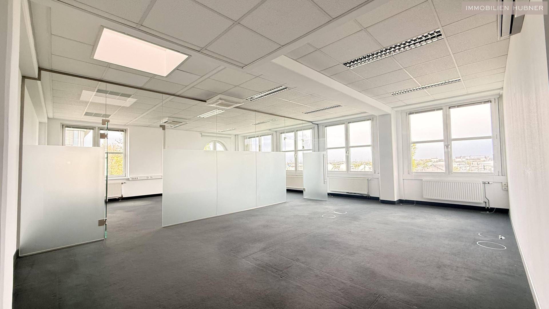 Großes, offenes Büro mit Teppichboden, Glasabtrennungen und vielen Fenstern, die eine moderne Arbeitsumgebung schaffen.