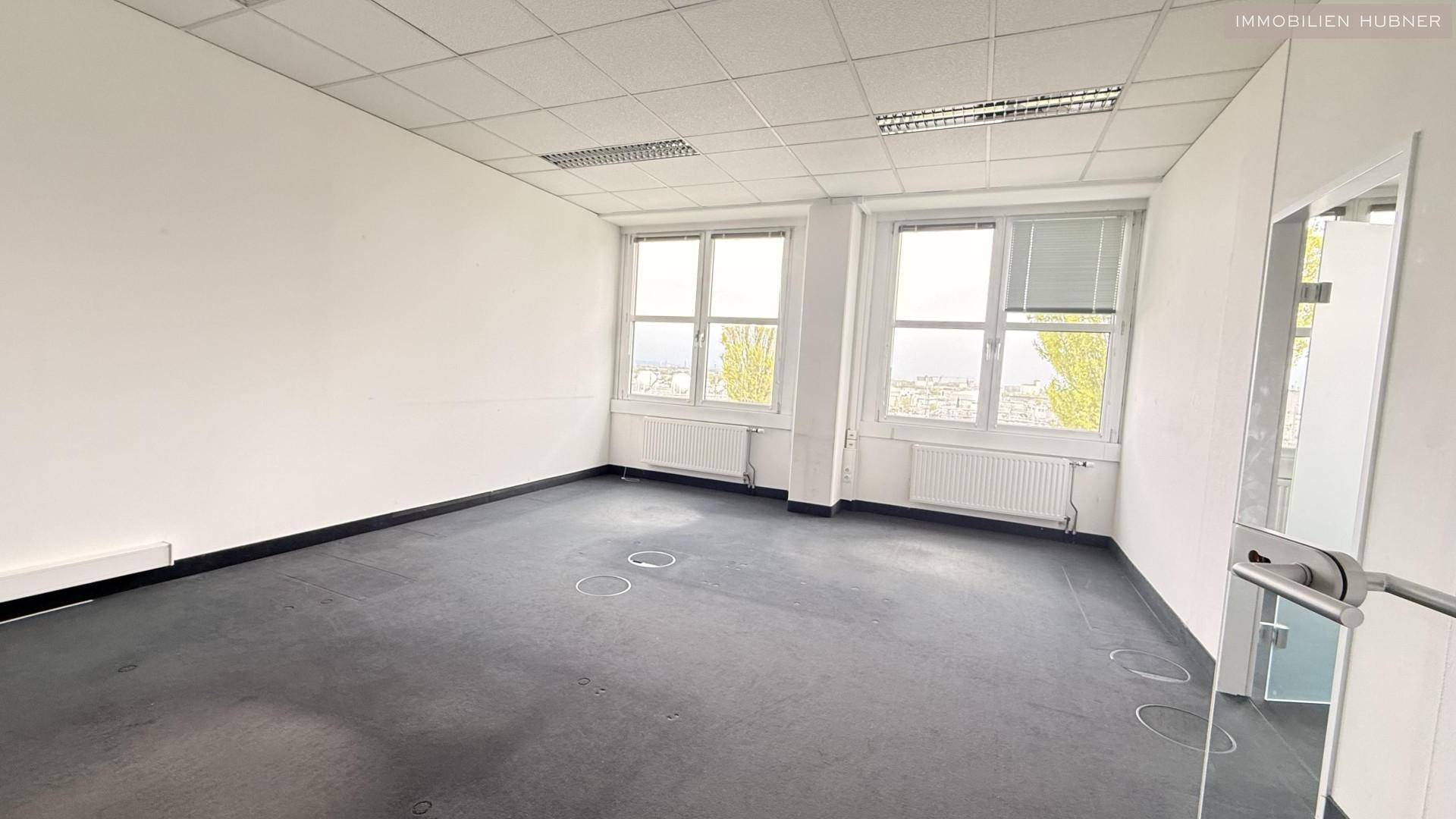 Helles Büro mit Teppichboden und zwei großen Fenstern, die viel Tageslicht hereinlassen und eine weite Aussicht bieten.