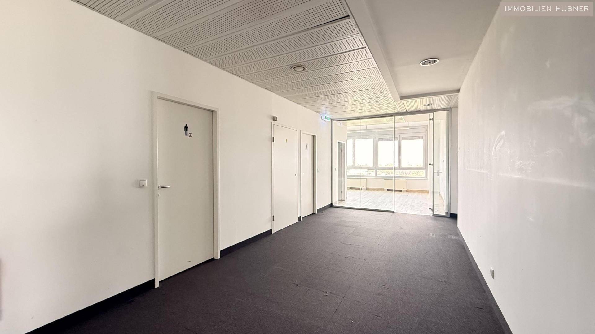 Langer Flur mit Teppichboden, abgehängter Decke und Zugang zu mehreren Büros, gut beleuchtet.
