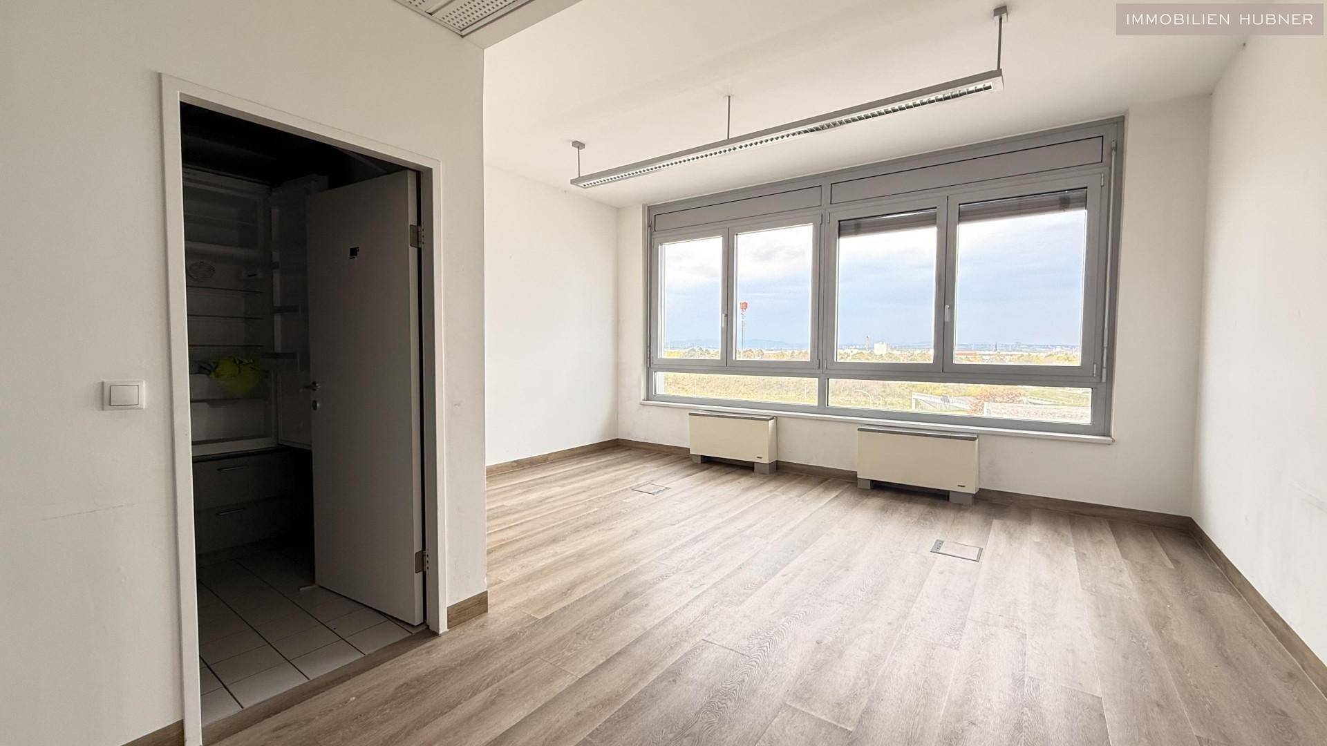 Büroraum mit hellem Laminatboden, großen Fenstern und Zugang zu einem Abstellraum, bietet vielseitige Nutzungsmöglichkeiten.