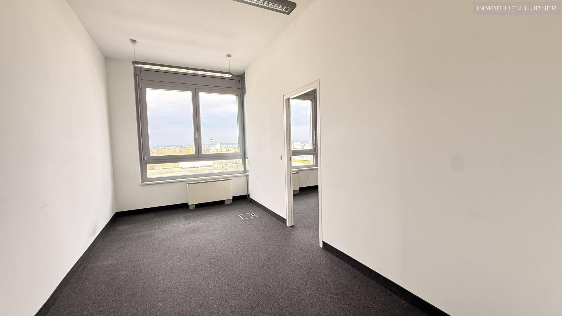 Büroraum mit Teppichboden, großen Fenstern und Zugang zu einem weiteren Raum, ideal für Teamarbeit.