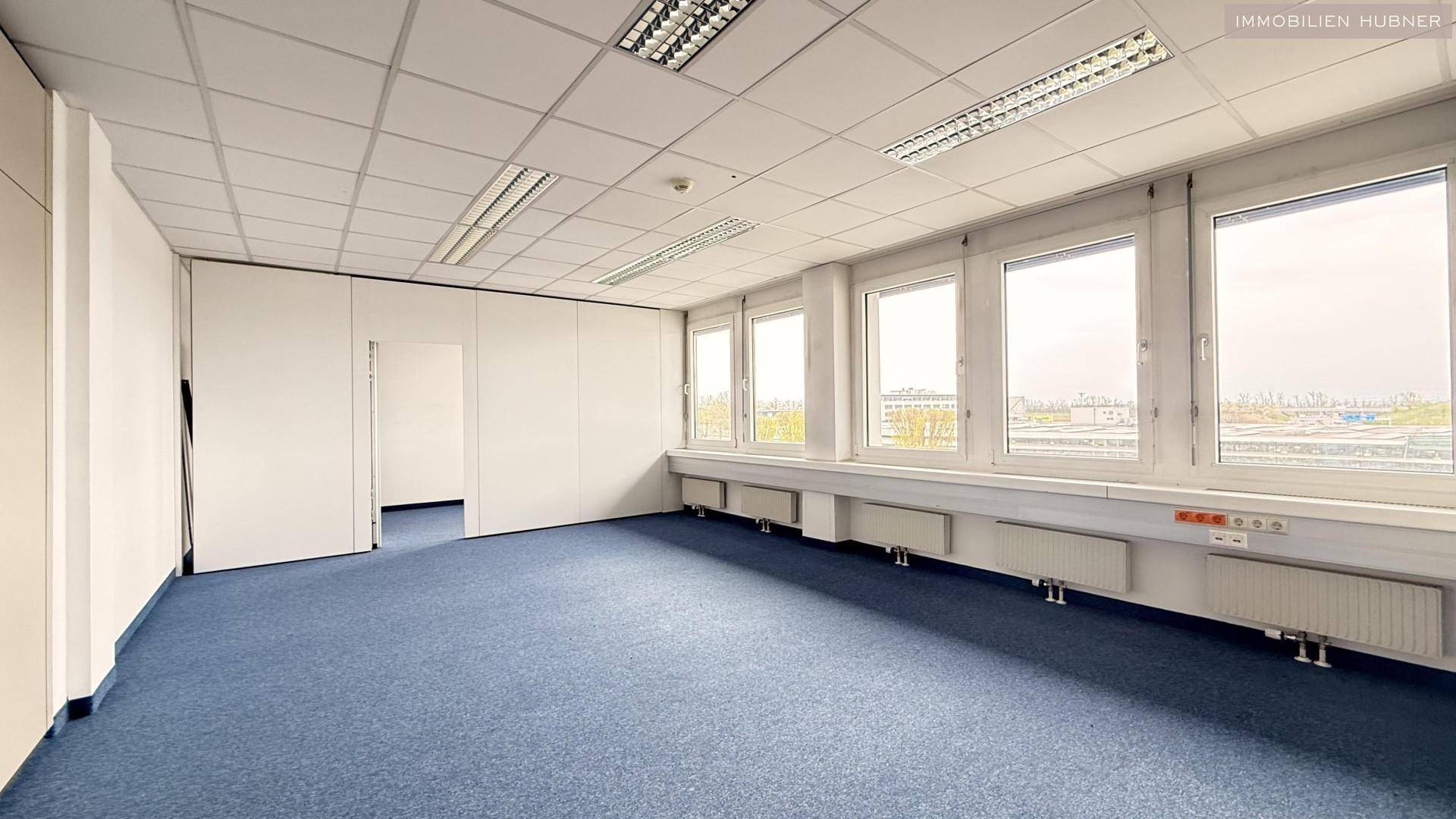 Geräumiges Büro mit blauer Teppichboden, modularen Wänden und Fenstern mit Ausblick.