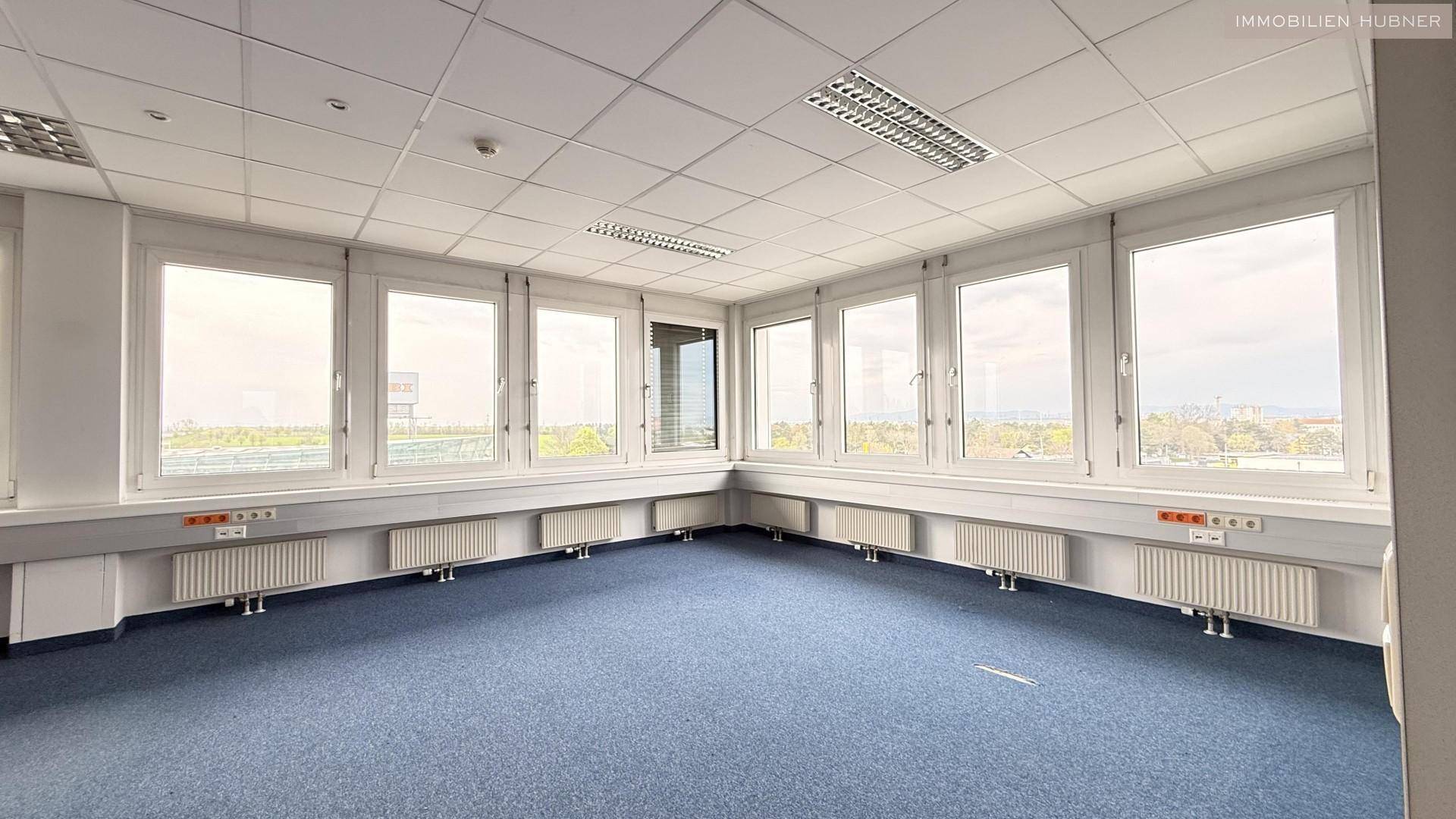 Großes Eckbüro mit vielen Fenstern, blauer Teppichboden und weitem Ausblick.