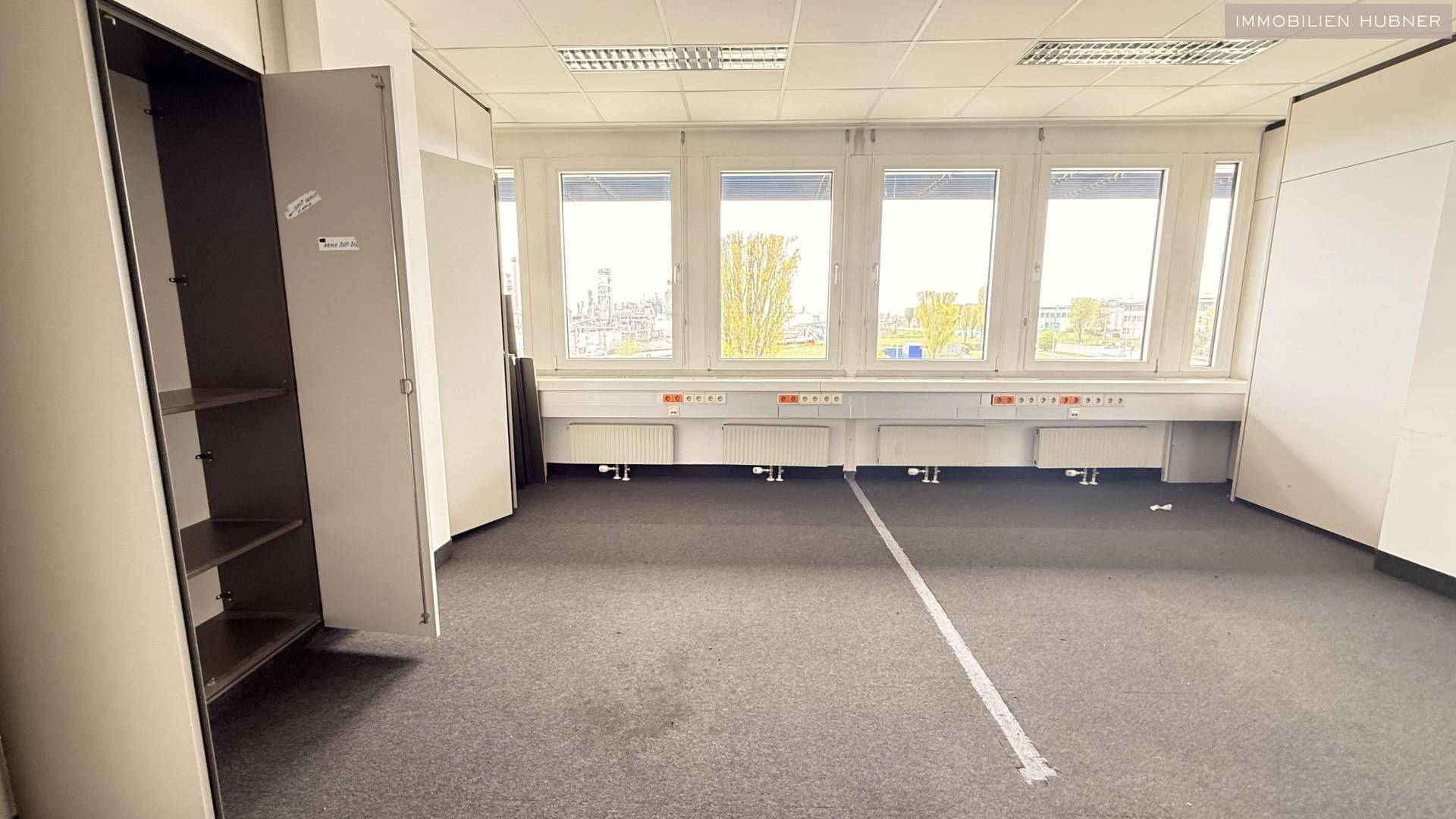 Leeres Büro mit Teppichboden, Einbauschrank und großen Fenstern, die viel Tageslicht hereinlassen.