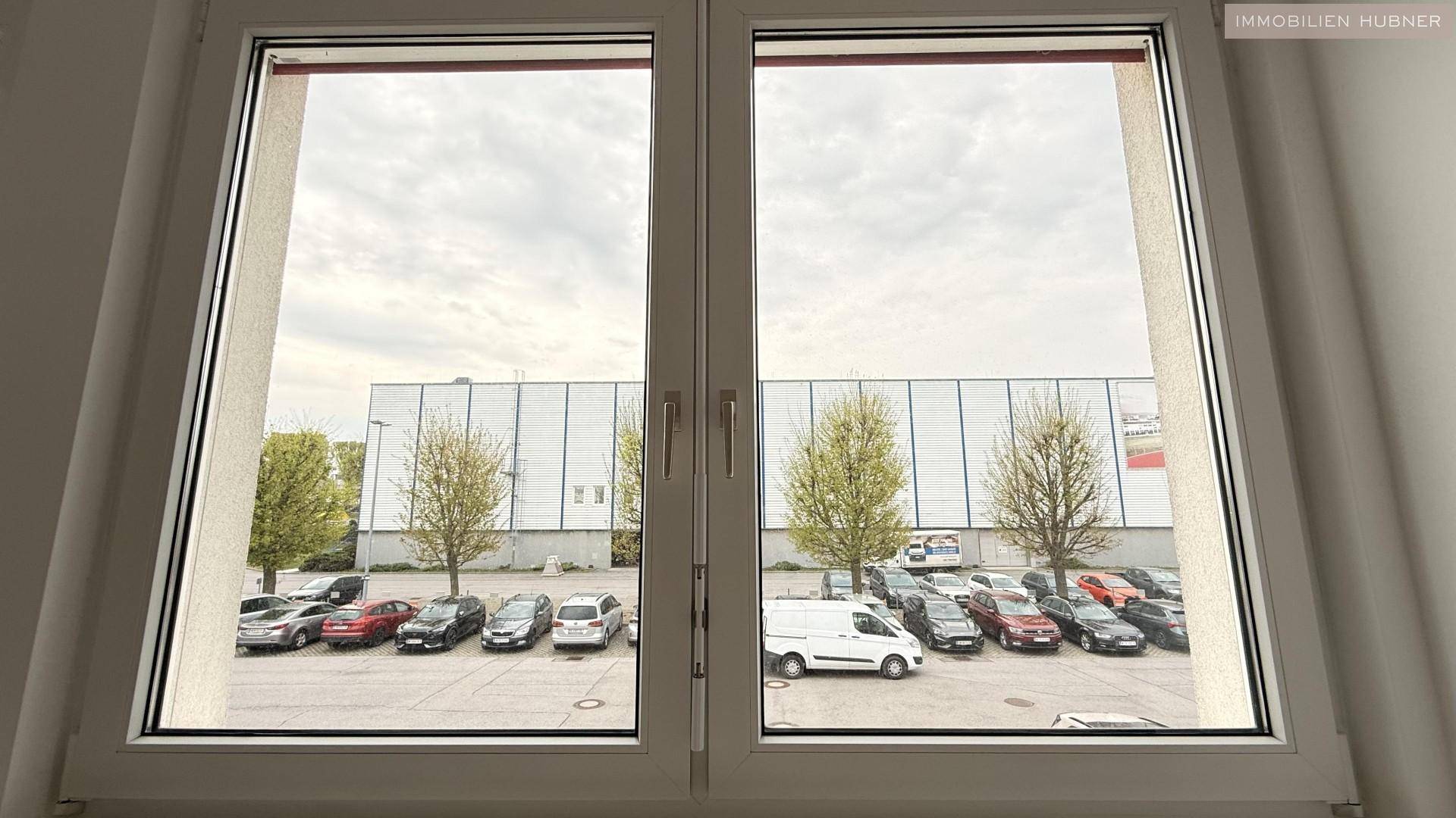 Blick aus dem Fenster eines Büros auf einen Parkplatz und ein Industriegebäude unter bewölktem Himmel.