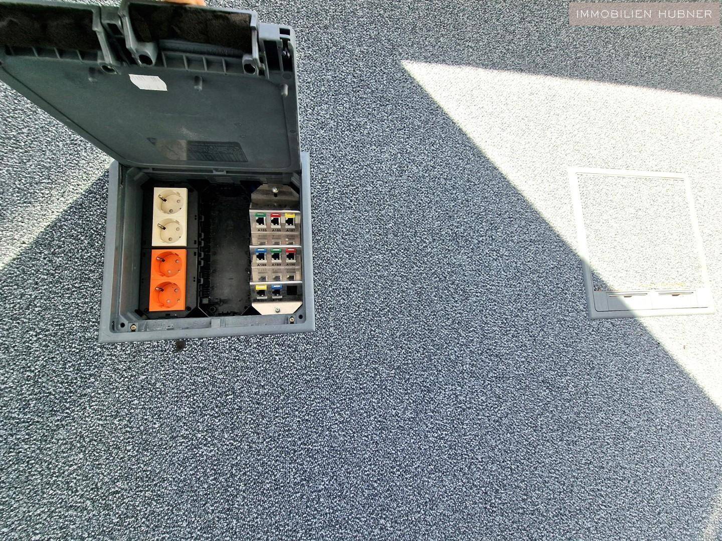 Offener Bodenkasten mit elektrischen Anschlüssen und Steckdosen auf einem Teppichboden.