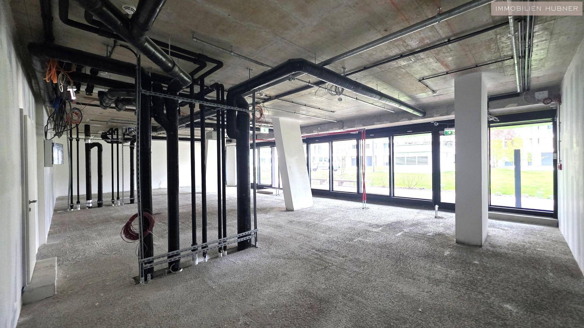 Geräumiger, unfertiger Büroraum mit sichtbaren Installationen an der Decke und großen Fenstern zur Außenseite.