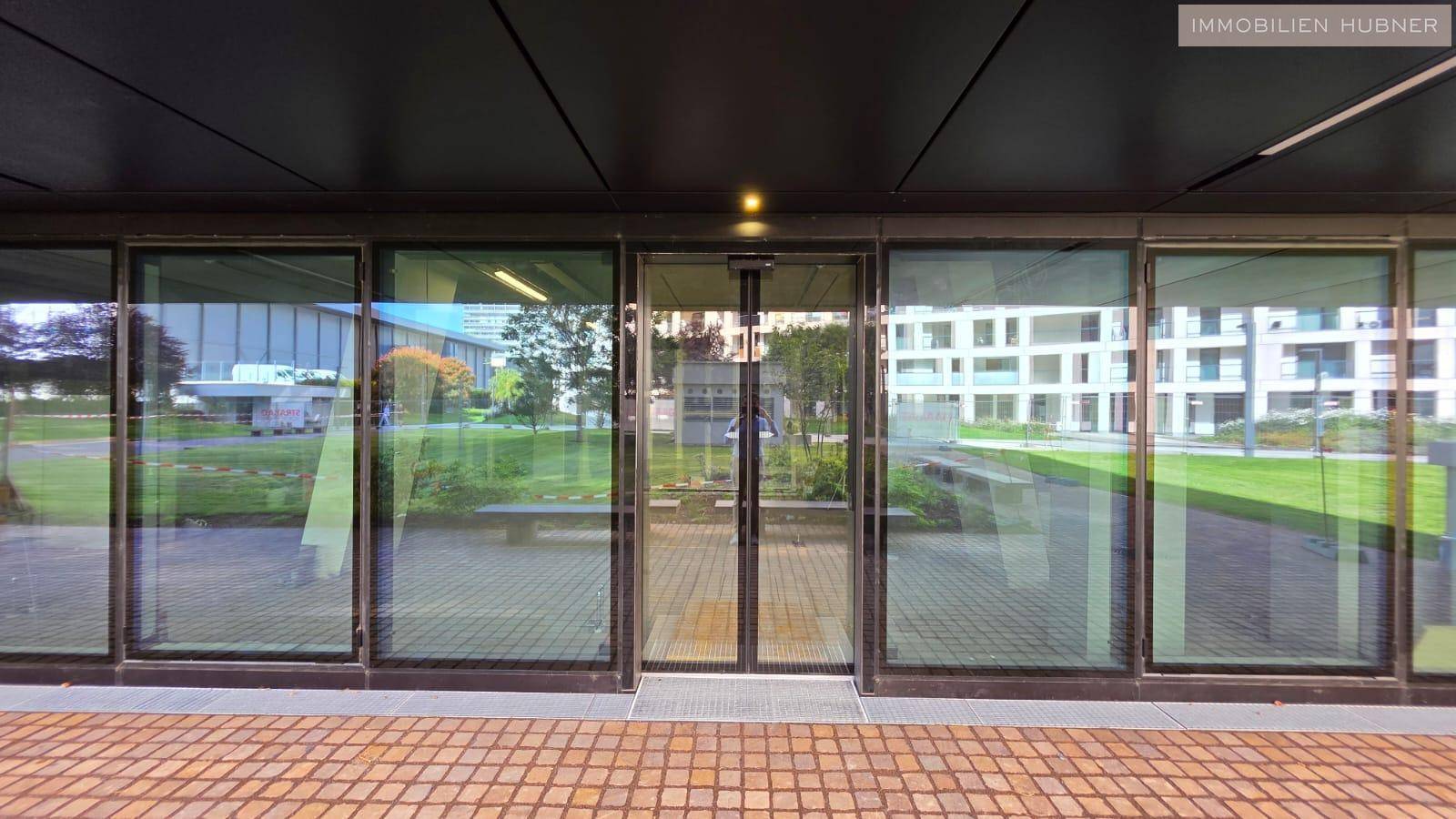 Moderner Gebäudeeingang mit großer Glasfront und Blick auf eine gepflasterte Fläche und Grünanlagen.