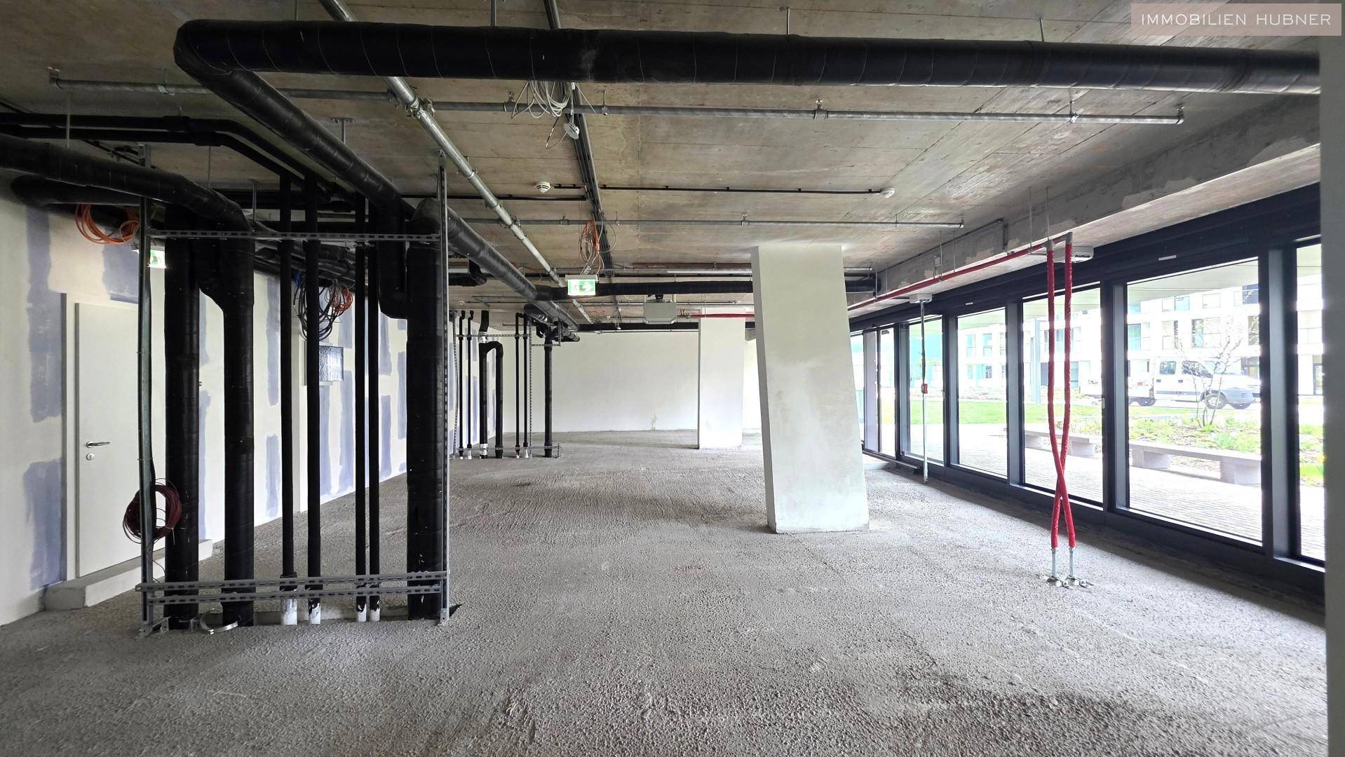 Weiterer Blick in den unfertigen Büroraum mit freiliegenden Rohren und viel Tageslicht durch die Fensterfront.