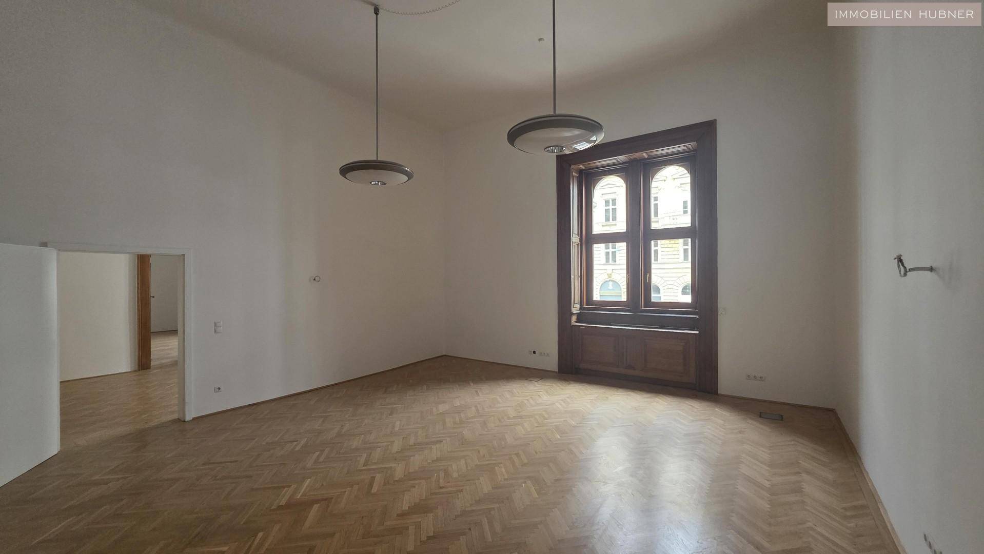 Geräumiger Raum mit hohen Decken, großen Fenstern und glänzendem Fischgrätparkettboden.