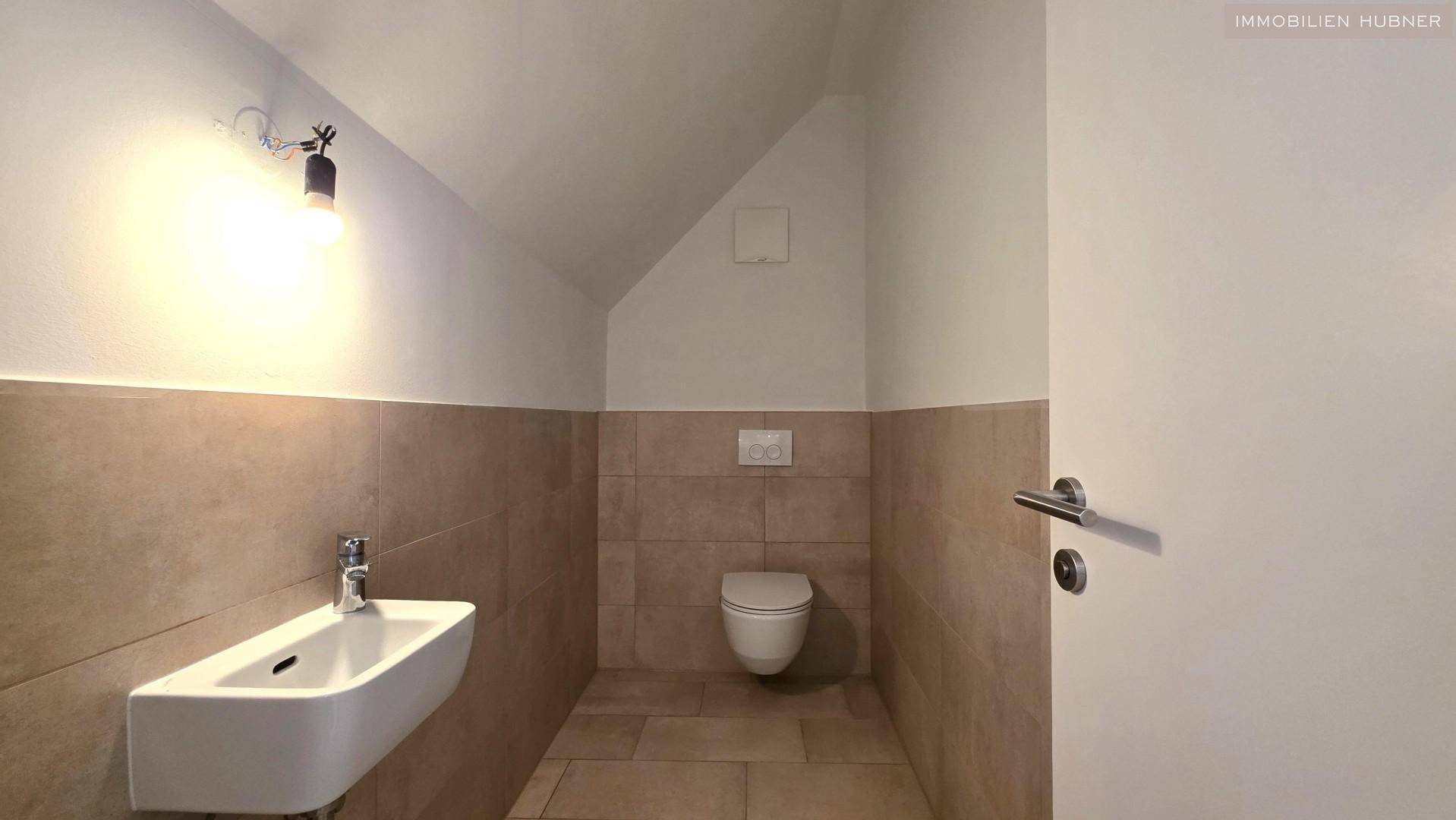 Gäste-WC mit wandhängender Toilette, kleinem Waschbecken und beigefarbenen Fliesen.