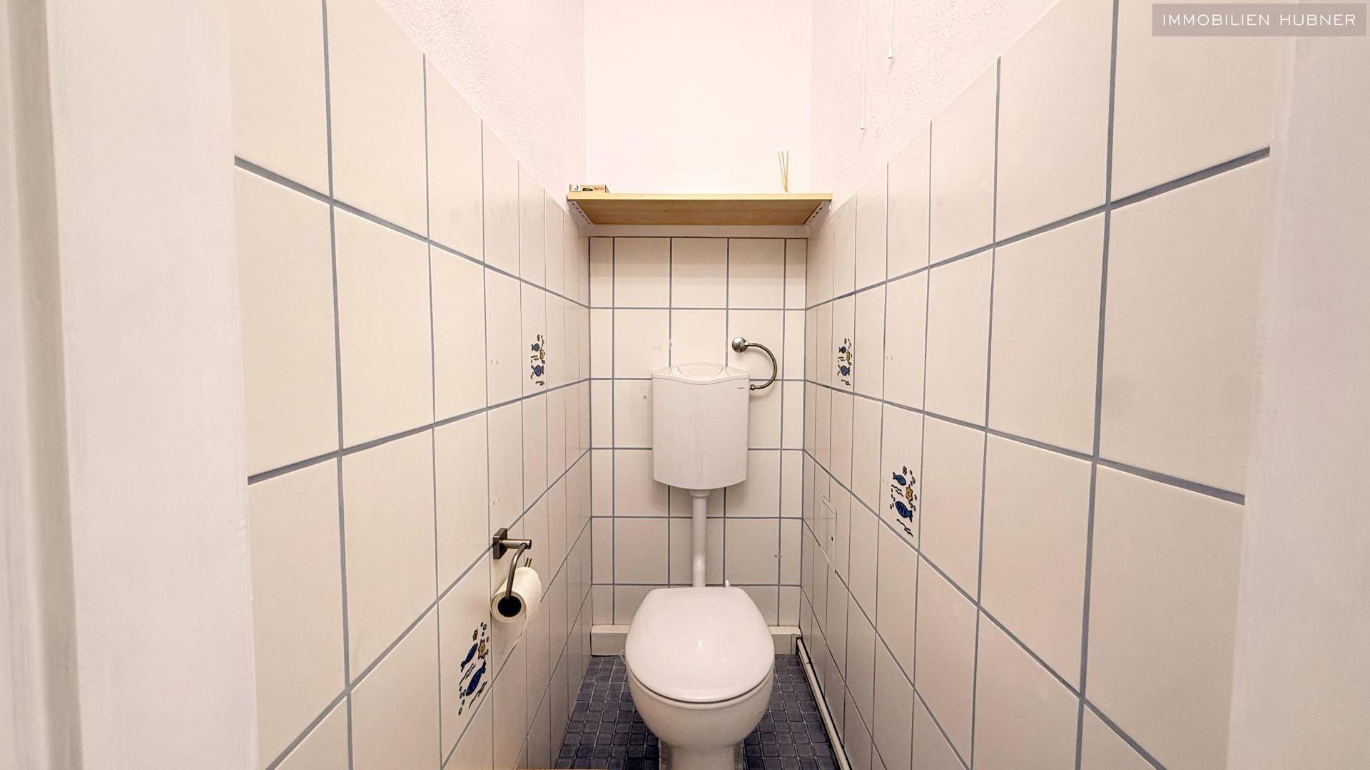 Separate Toilette mit weißen Fliesen und einem kleinen Regal, praktisch und sauber.