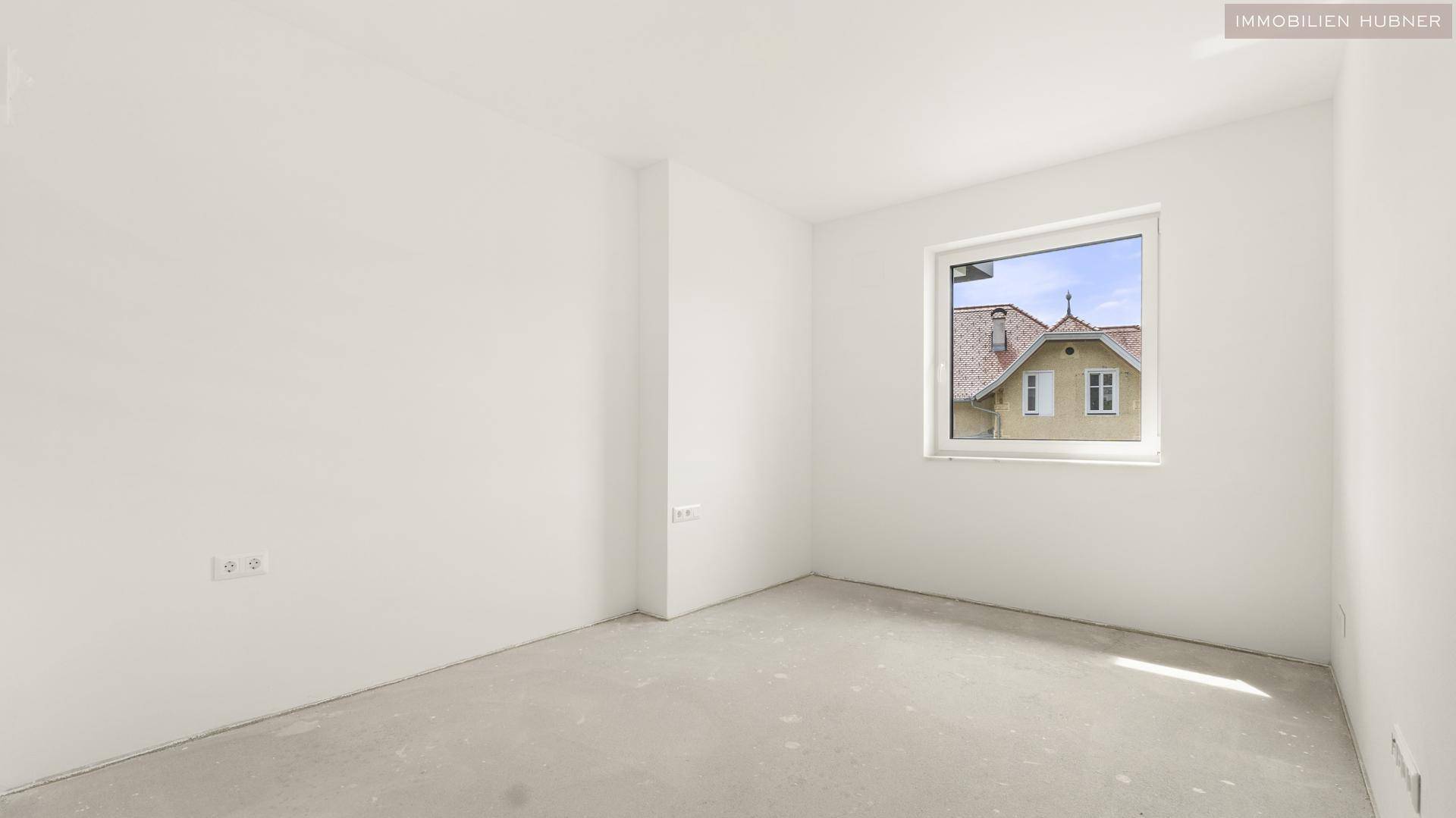 Leeres Zimmer mit weiß gestrichenen Wänden und einem Fenster mit Blick auf ein Nachbarhaus.