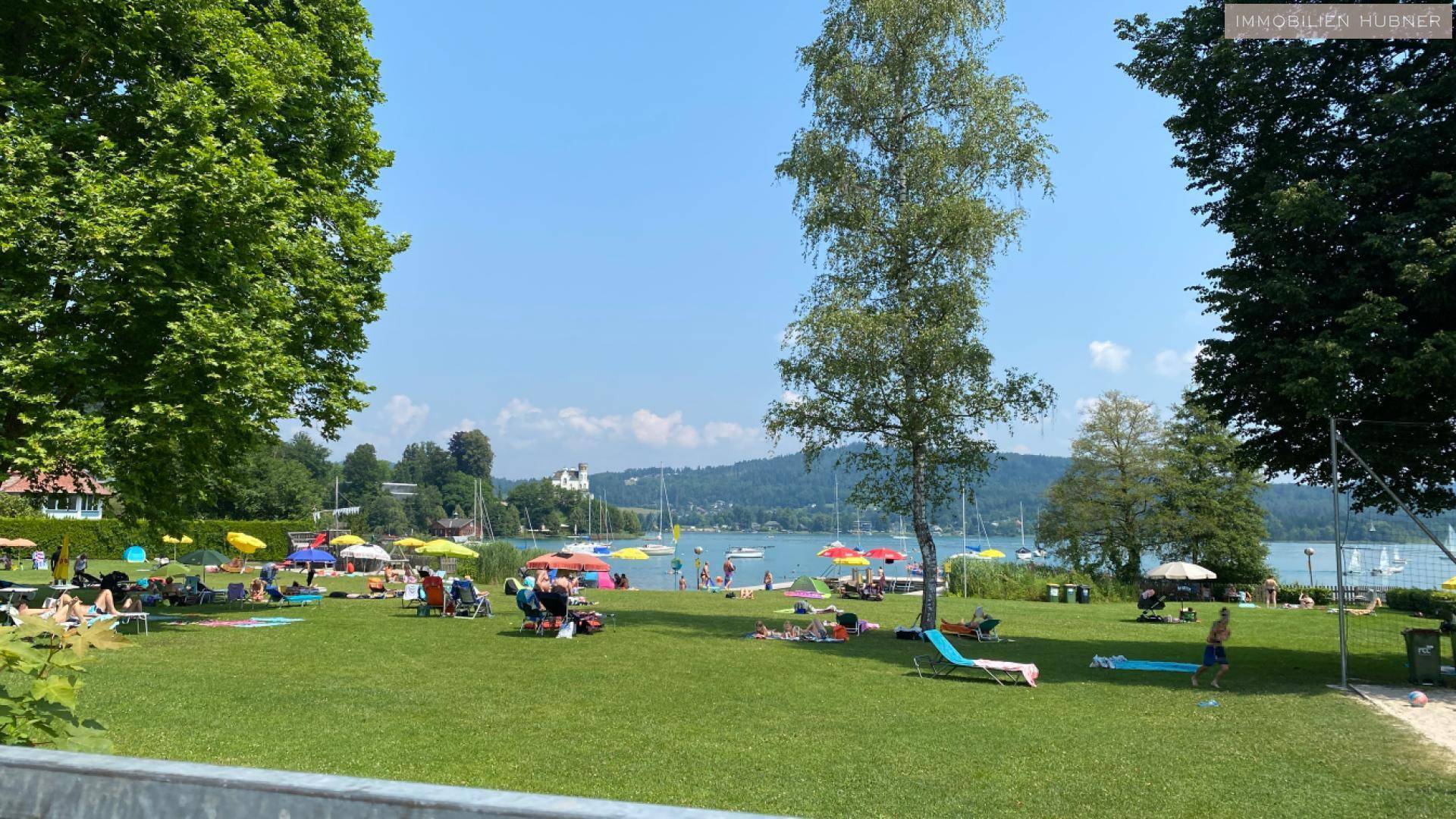 Sonniger Badestrand am See mit vielen Menschen, Bäumen und Booten im Hintergrund.