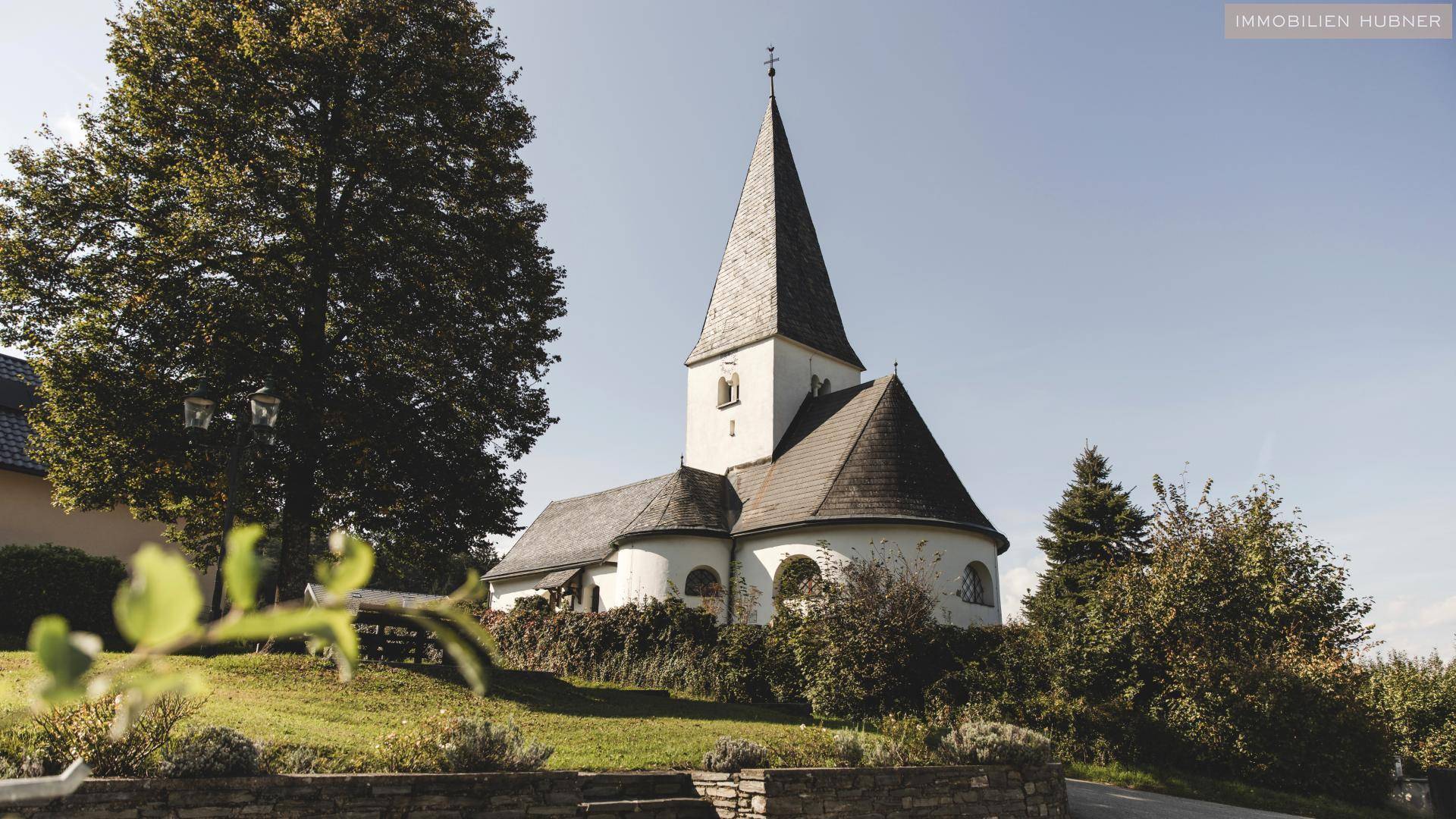 Historische Kirche mit spitzem Turm, umgeben von Bäumen und grüner Landschaft unter klarem Himmel.