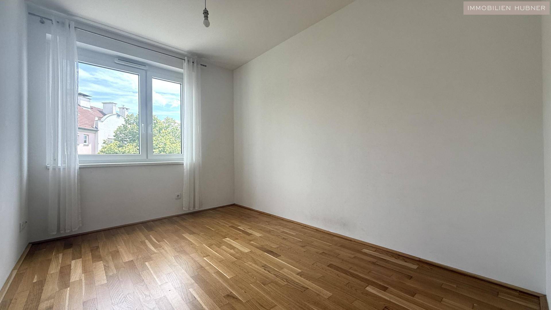 Helles Schlafzimmer mit Parkettboden und großem Fenster, das viel Tageslicht hereinlässt.