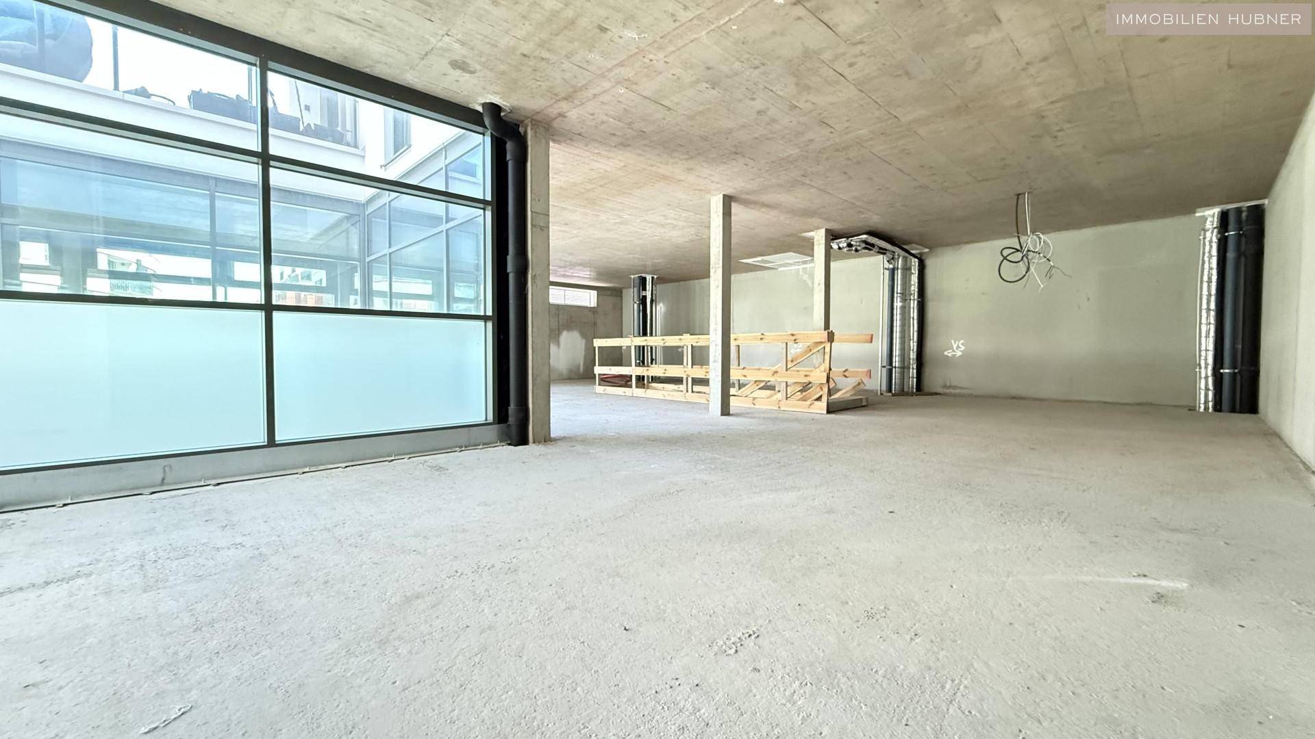 Unfertiger Gewerberaum mit großen Fenstern und sichtbaren Deckeninstallationen, bereit für den Ausbau.