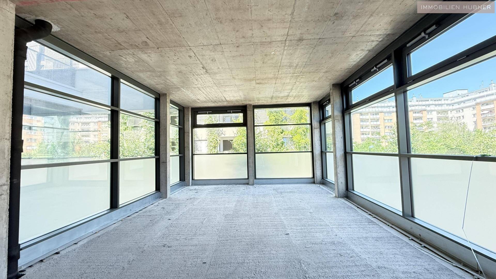 Langer, heller Gewerberaum im Rohbau mit umlaufenden Fensterfronten und Betonboden.
