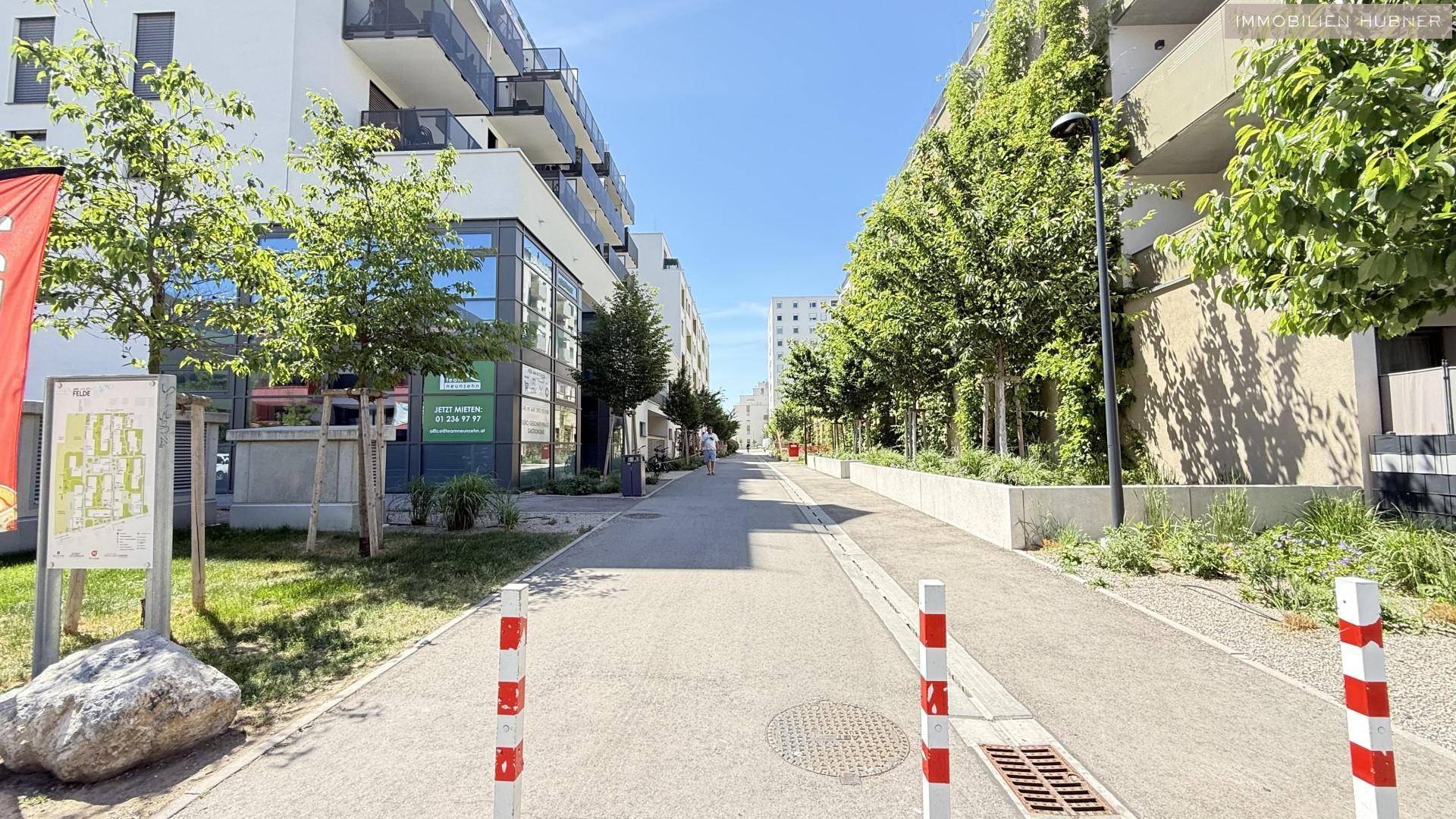 Fußgängerzone zwischen modernen Gebäuden mit viel Grün und Bäumen an einem sonnigen Tag.