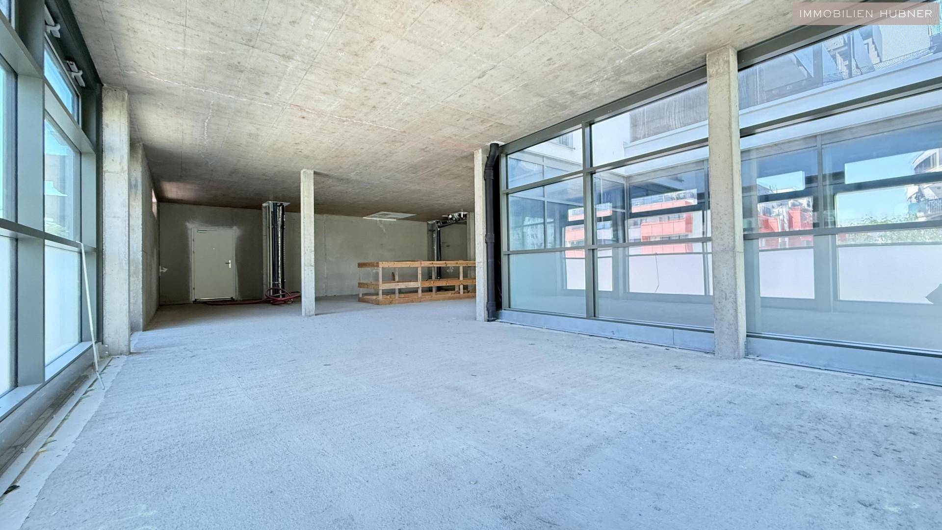Heller Rohbau-Gewerberaum mit Betonboden und großen Fensterflächen, die viel Tageslicht bieten.