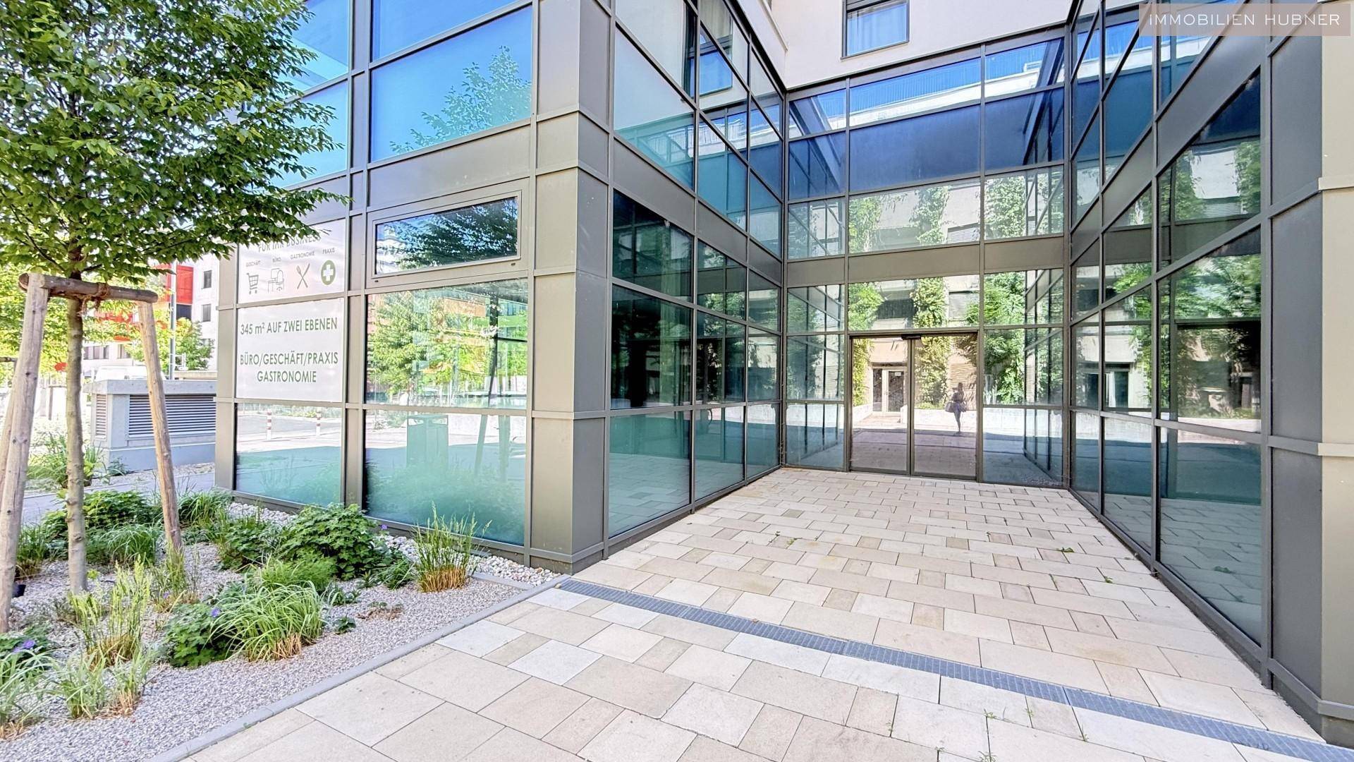 Eingangsbereich eines modernen Gebäudes mit großer Glasfront und gepflastertem Vorplatz.