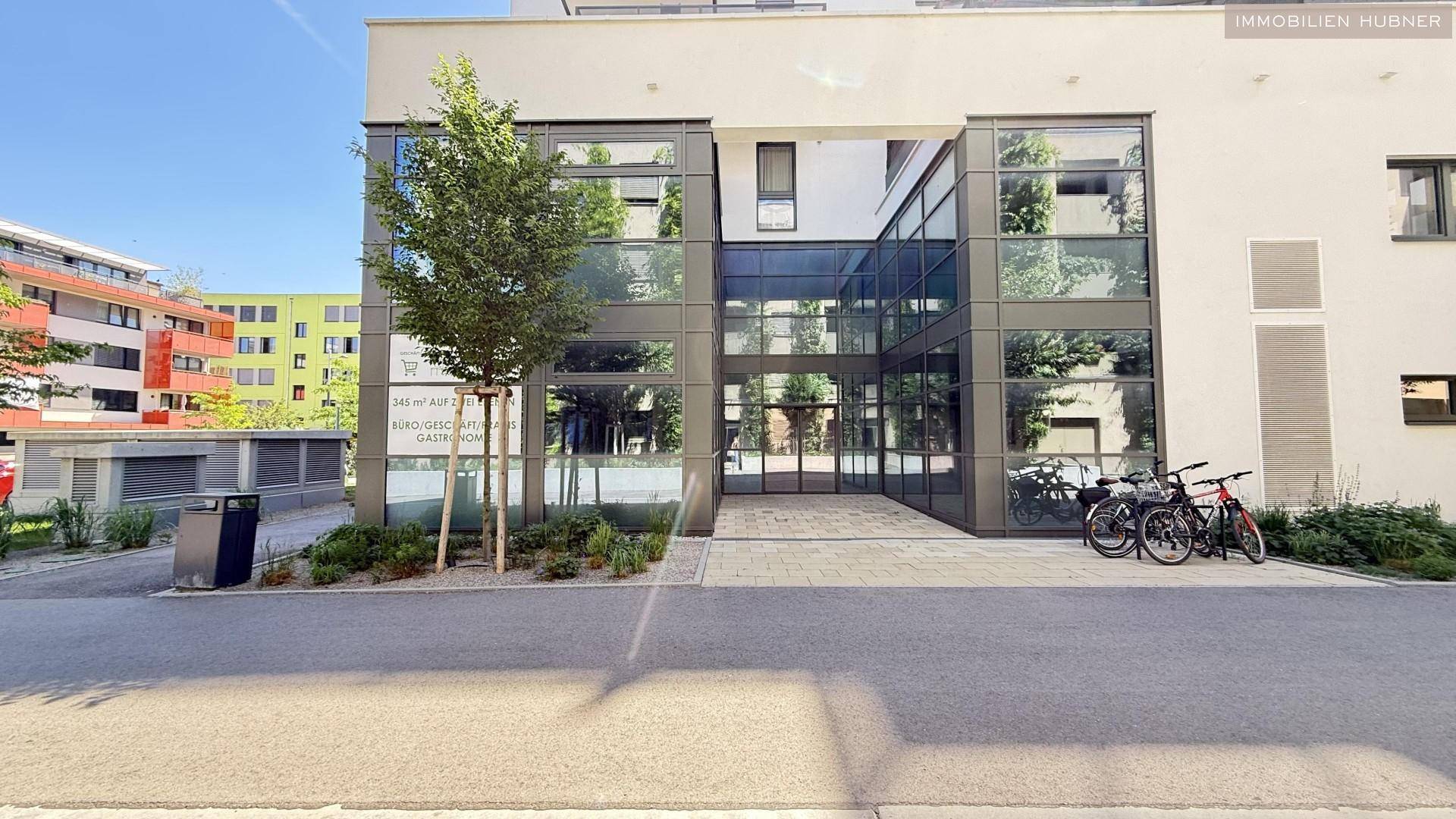 Eingangsbereich eines modernen Gebäudes mit großer Glasfront und gepflastertem Zugang, Fahrräder abgestellt.