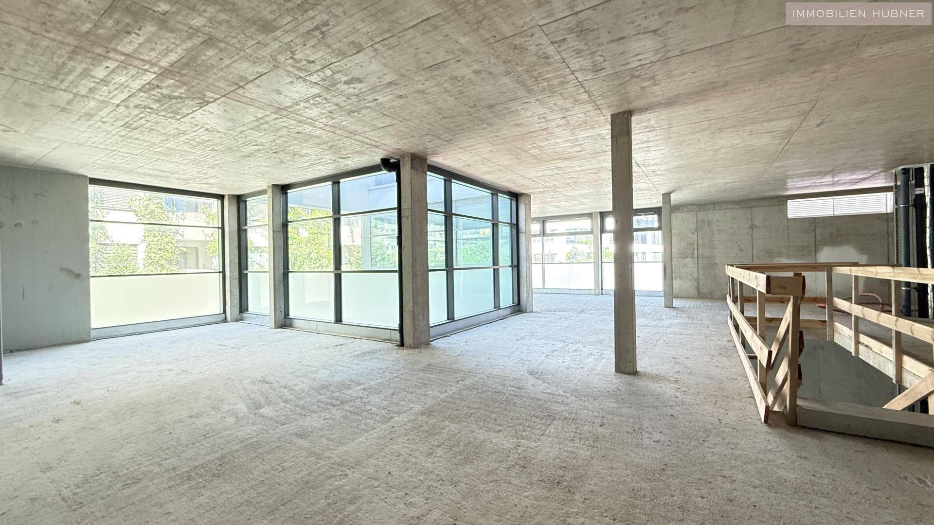 Großer, heller Gewerberaum im Rohbau mit Betonstützen und umlaufenden Fensterfronten.