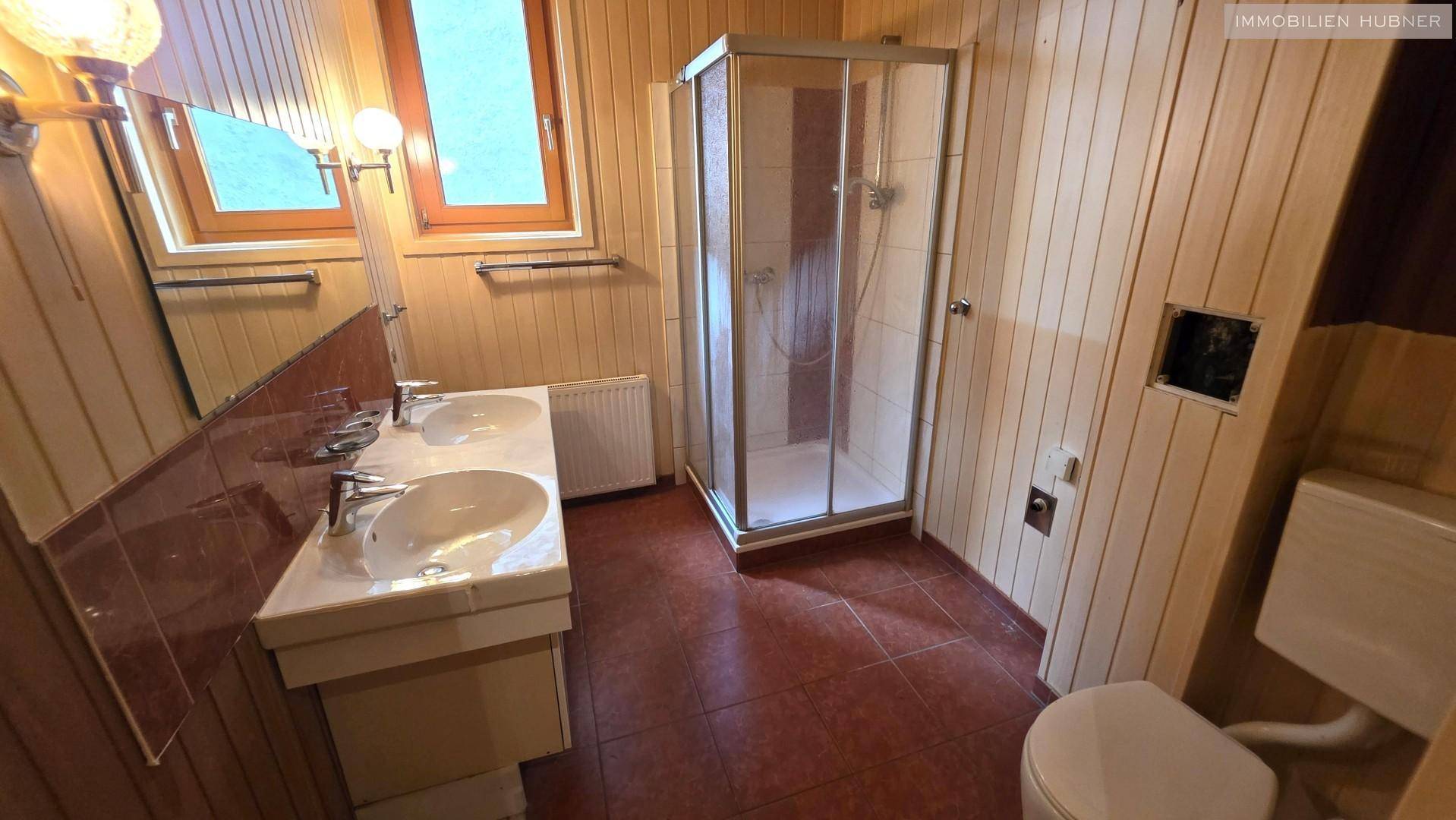 Badezimmer mit zwei Waschbecken, Duschkabine und Holzverkleidung, in älterem Zustand.