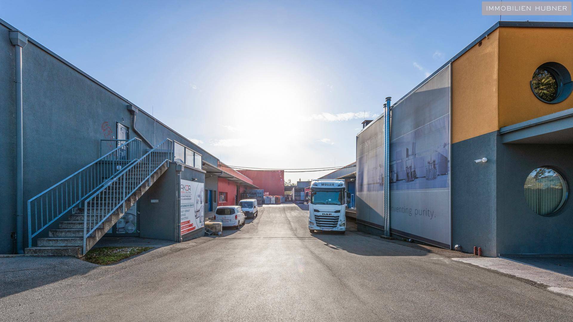 Zufahrt zwischen Industriegebäuden mit LKWs und einer Außentreppe unter blauem Himmel.