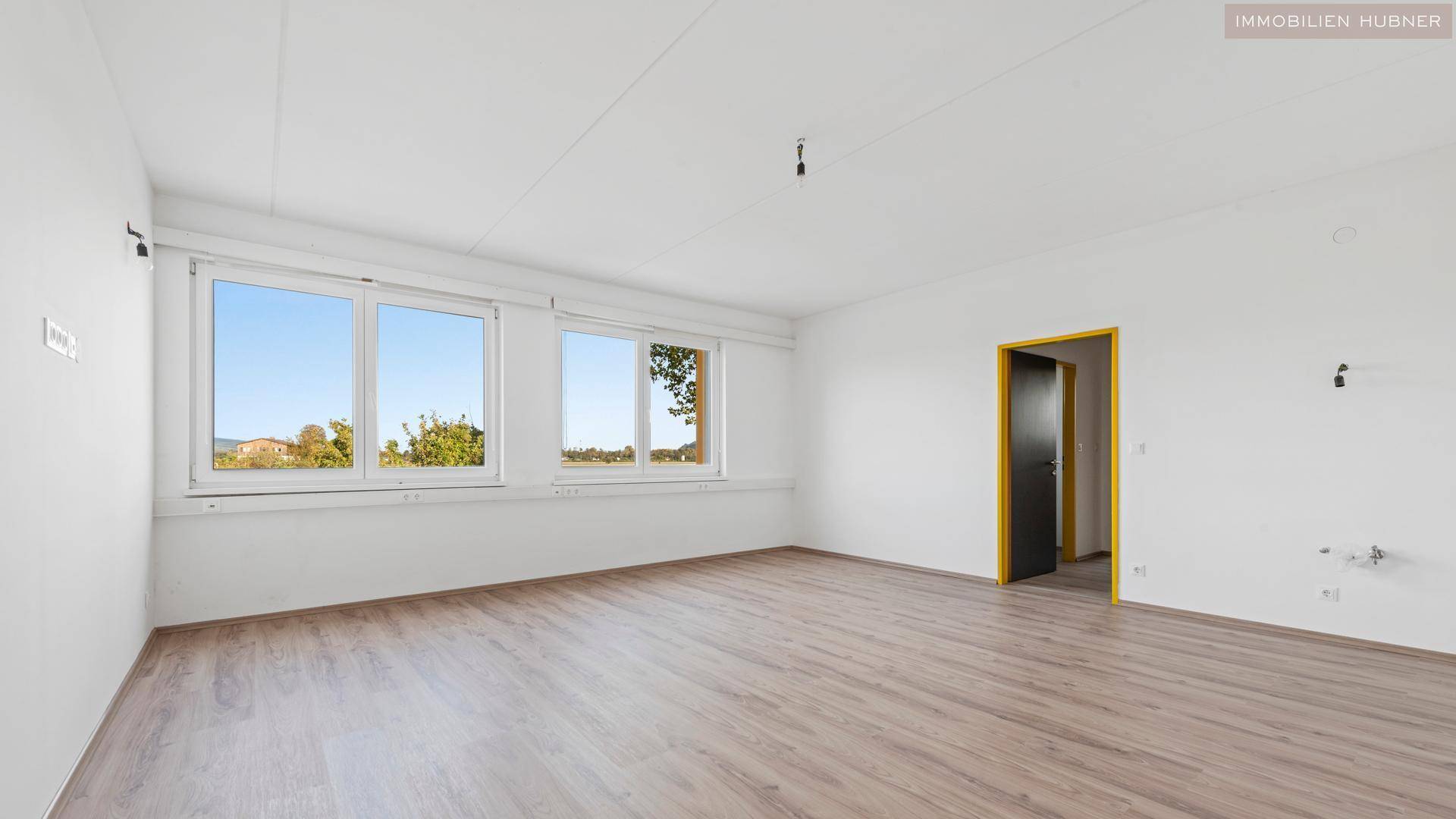 Geräumiger, heller Raum mit weißen Wänden, Laminatboden und Fenstern mit Blick ins Grüne.