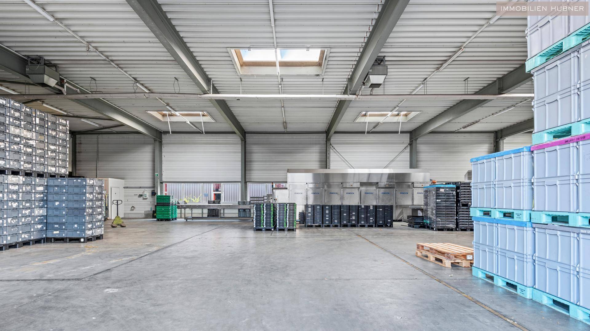 Weitläufige Lagerhalle mit hohen Decken, Oberlichtern und gestapelten Kisten auf dem Betonboden.