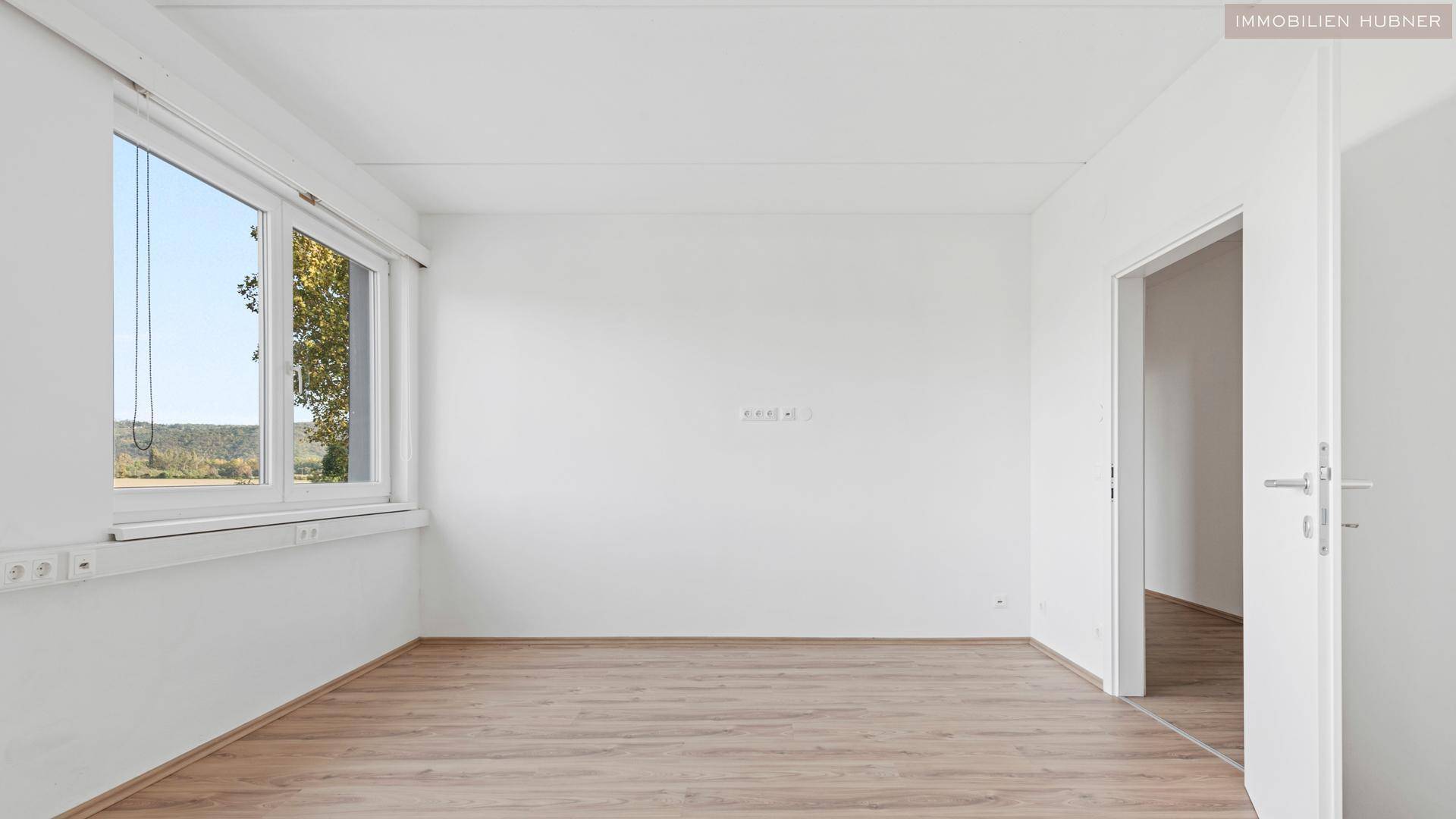Heller Raum mit weißen Wänden, Laminatboden und einem Fenster, das viel Tageslicht hereinlässt.