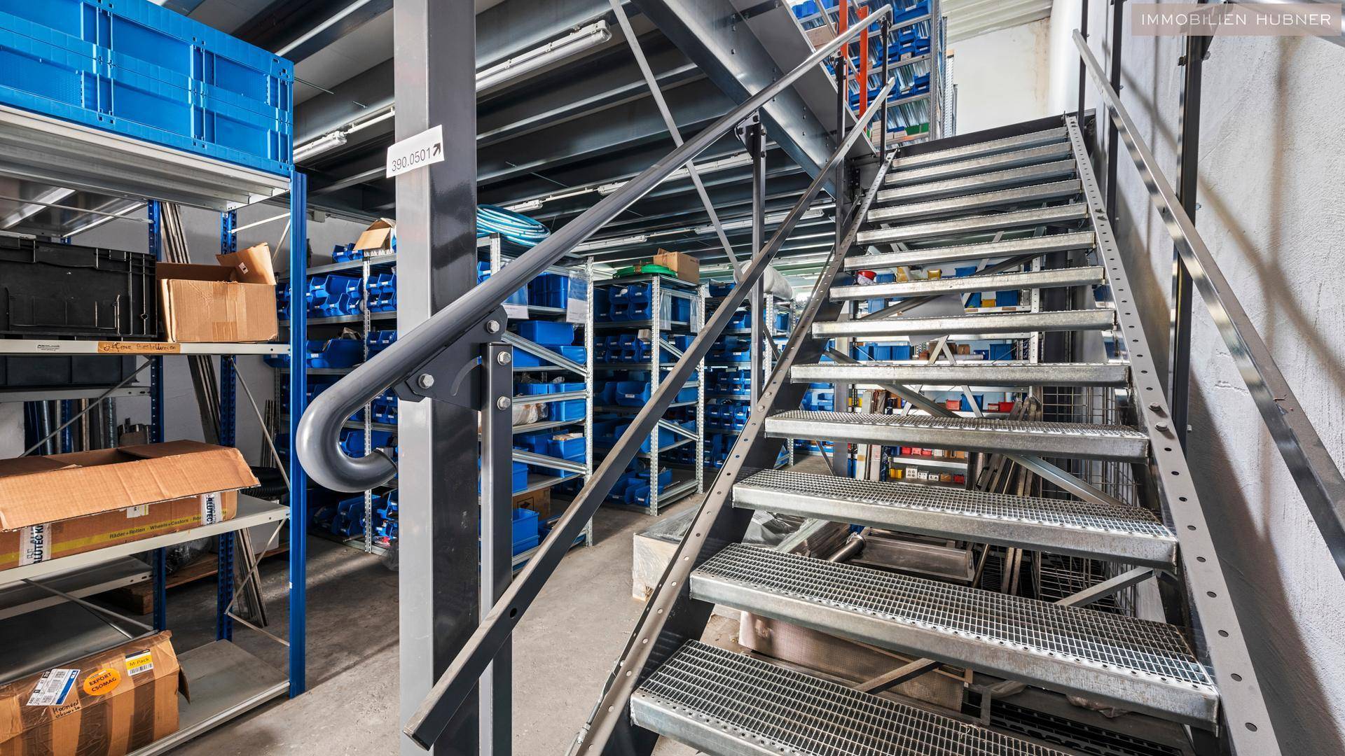 Metalltreppe in einem Lagerbereich mit Regalen voller blauer Kisten und verschiedenen Artikeln.