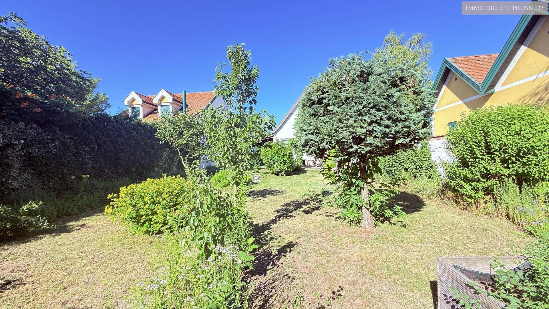 Großer Garten mit Rasen, jungen Bäumen und Blick auf benachbarte Häuser.