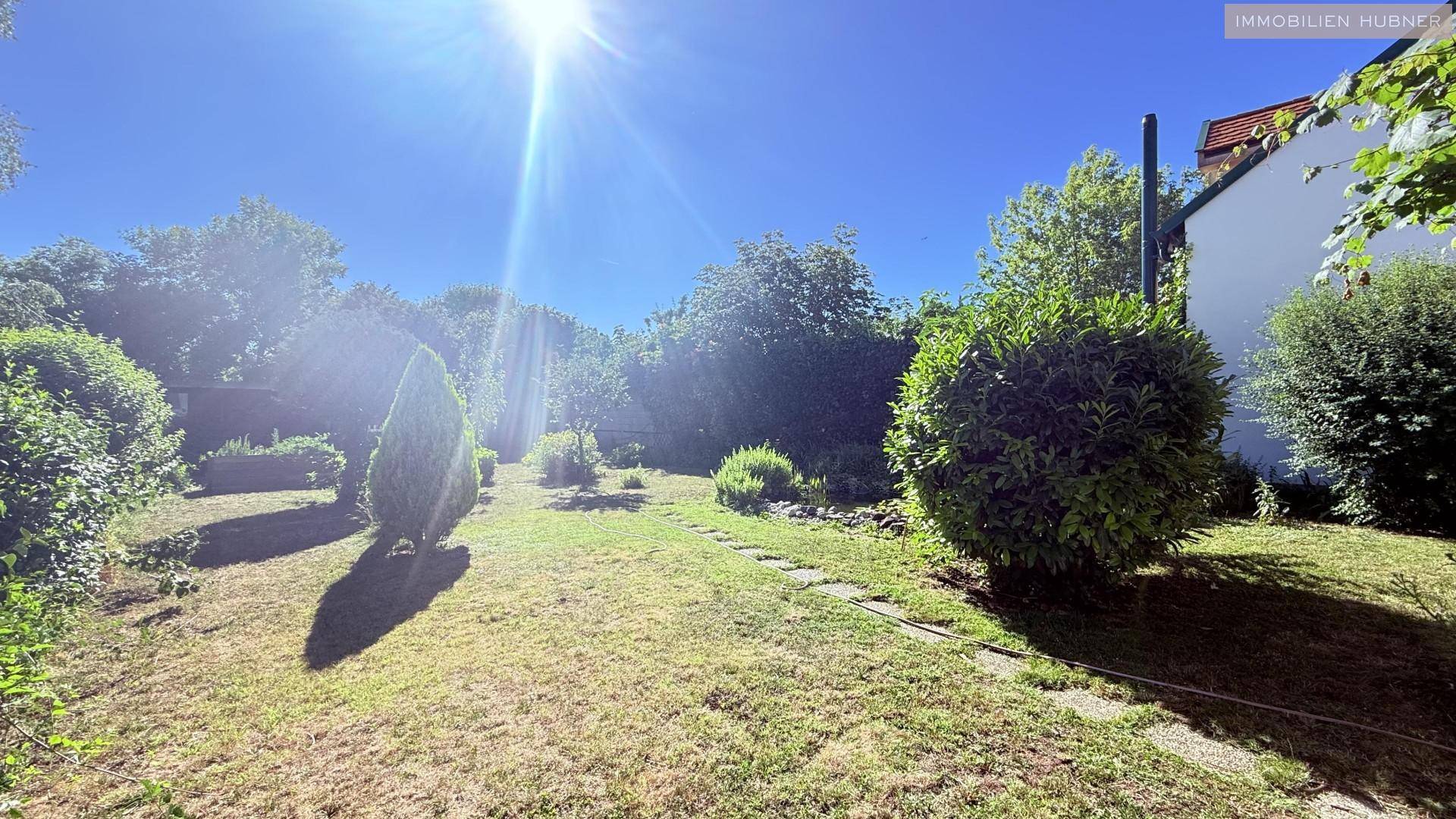 Weitläufiger Gartenbereich mit Rasenfläche, verschiedenen Bäumen und Sträuchern unter strahlendem Himmel.